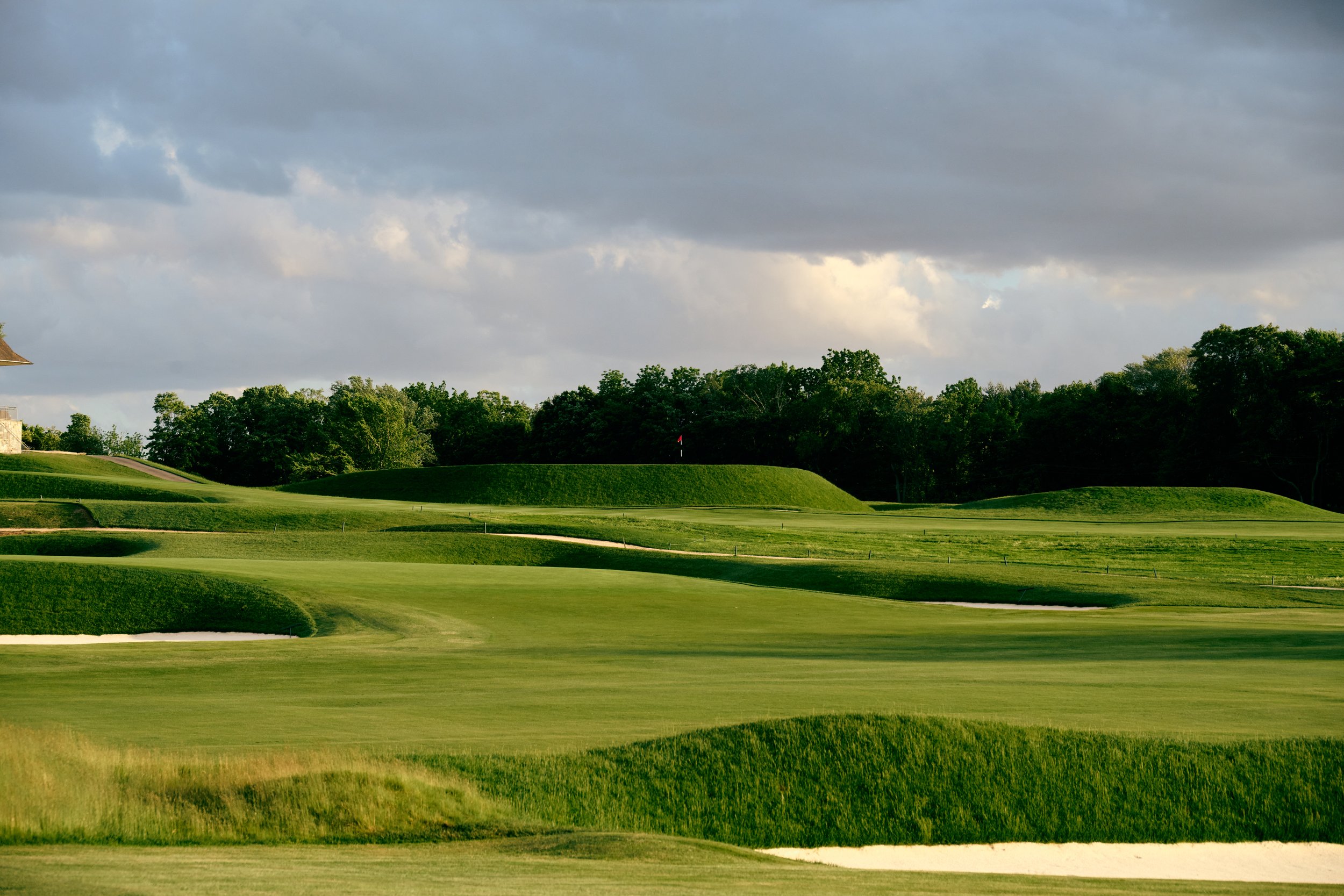 Scenic view of a golf course with lush green fairways, sand bunkers, a flag on a green, and trees in the background under a cloudy sky.