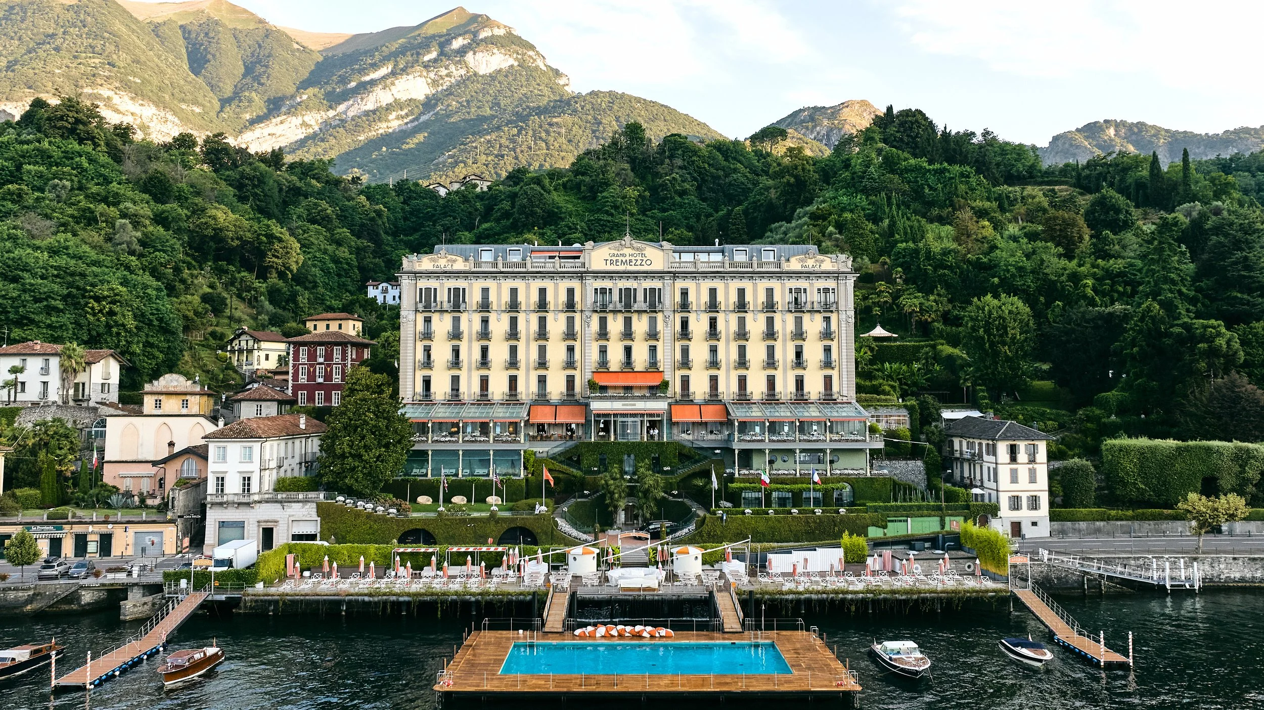 A large hotel building with yellow walls and black balconies, situated on a lake with boats and a dock in the foreground, lush green mountains in the background.