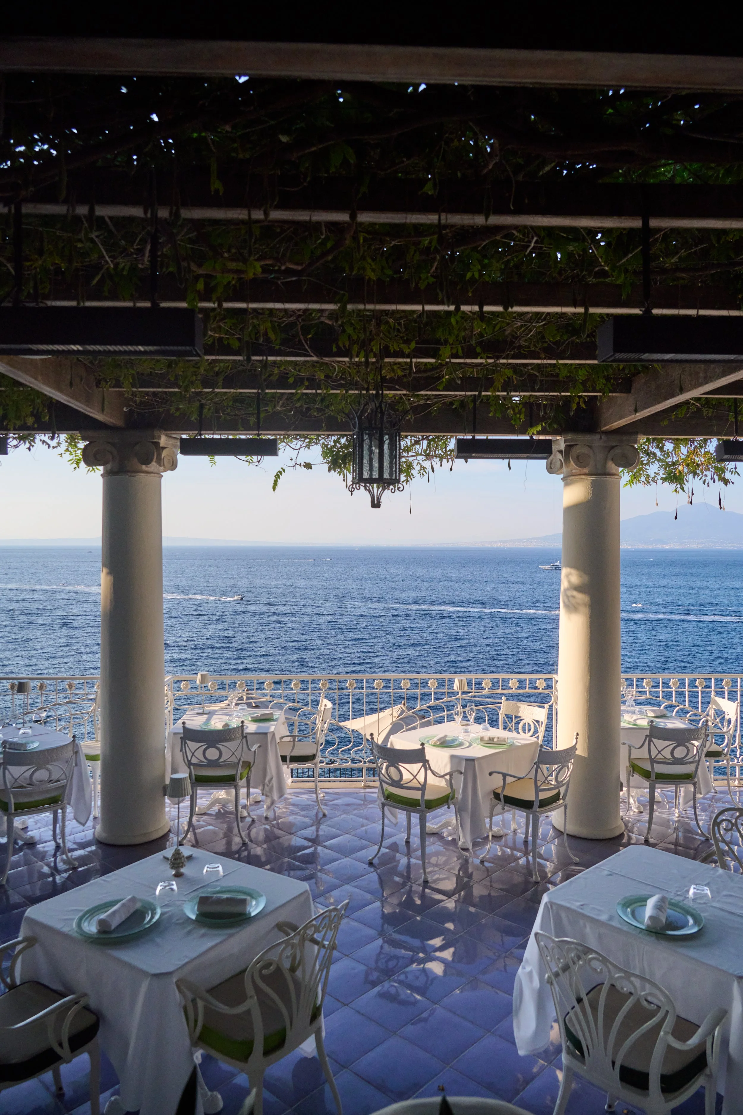 Outdoor dining area overlooking the ocean with several tables set with white tablecloths, plates, napkins, and silverware, framed by white columns and a wooden pergola.