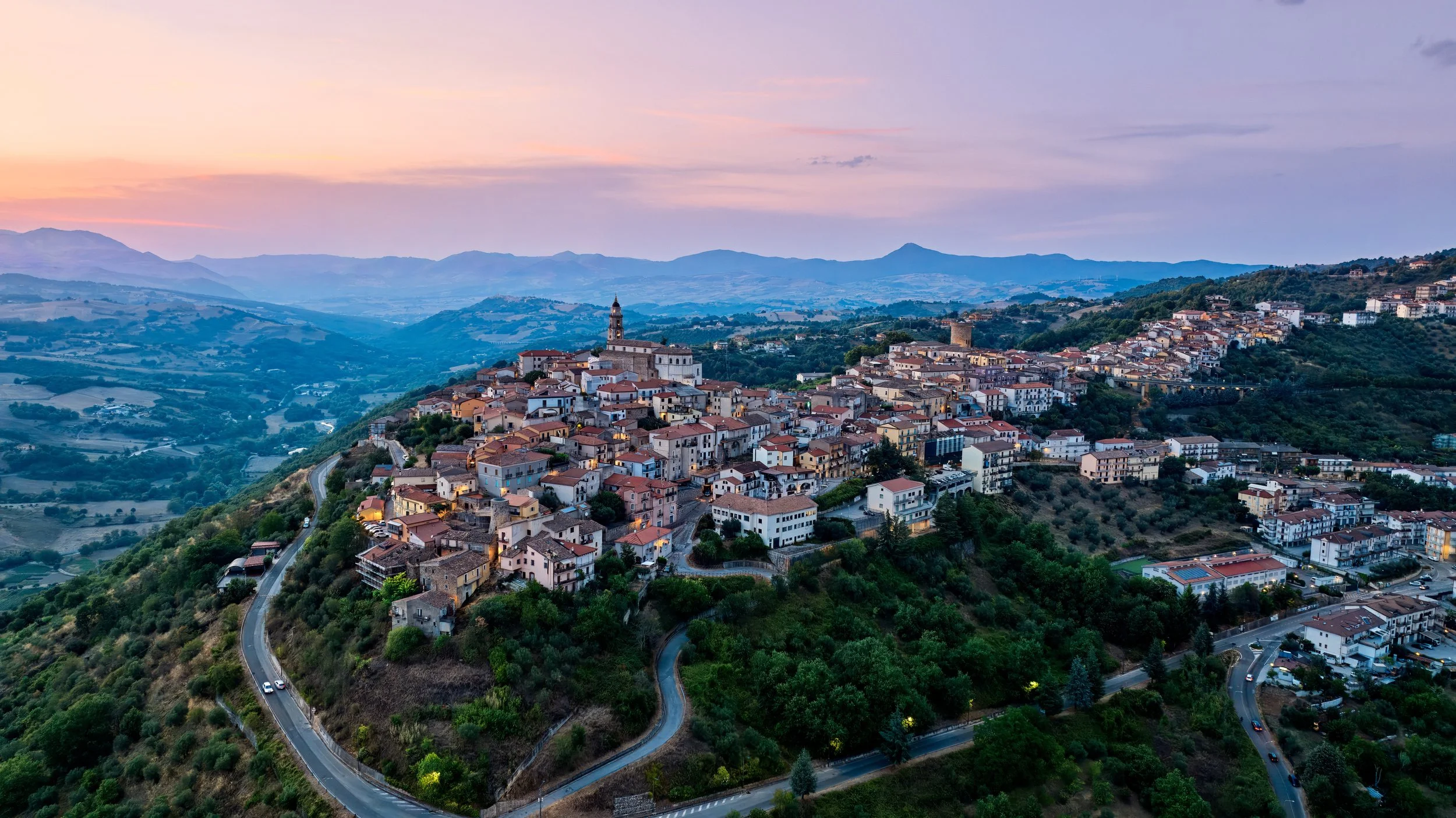 Aerial view of a hillside town at sunset, with narrow winding roads, Mediterranean-style houses with red-tiled roofs, a church tower, and lush green hills in the background.