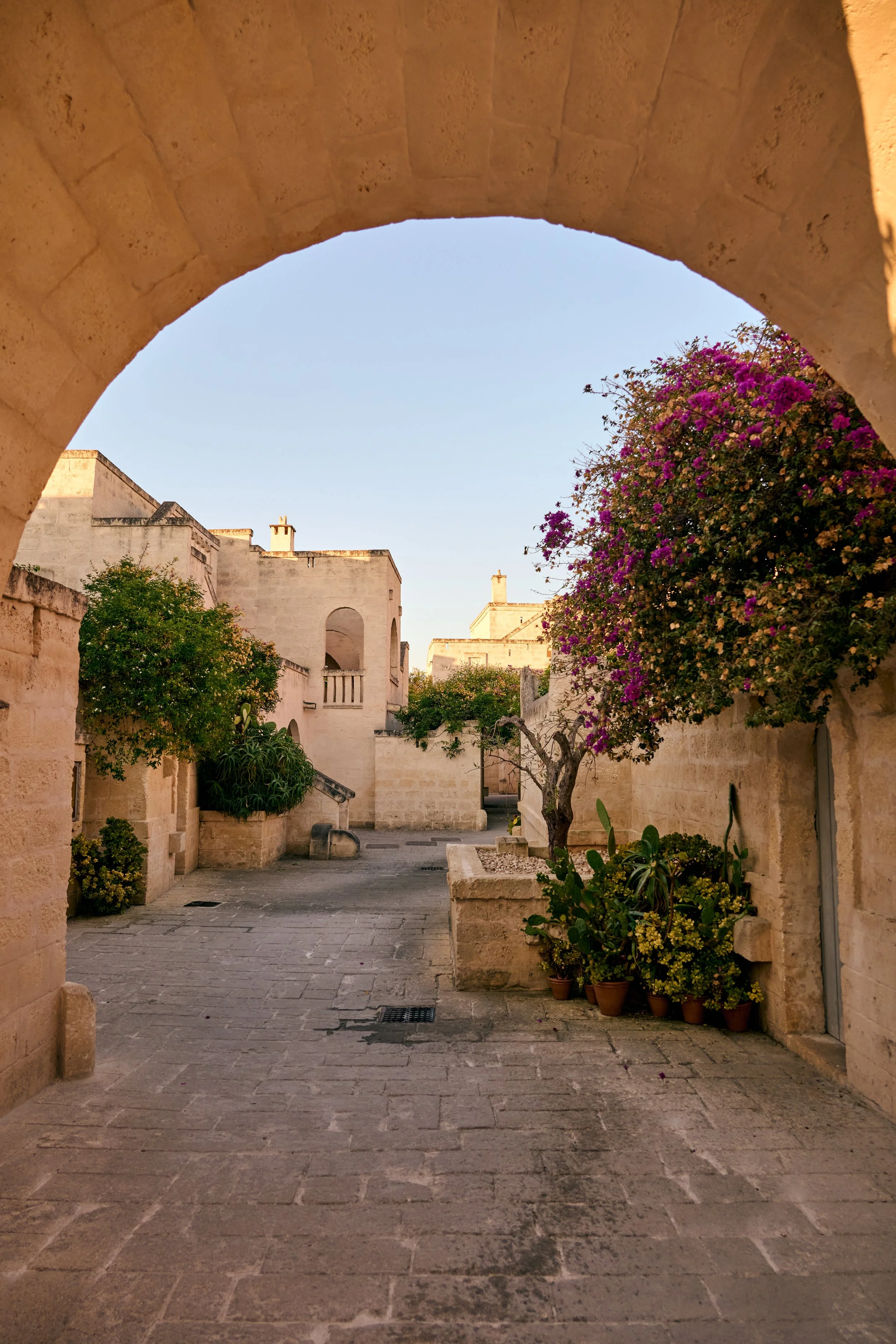 A view of a quiet street framed by an archway, with stone buildings, potted plants, and flowering trees in a Mediterranean or European town.