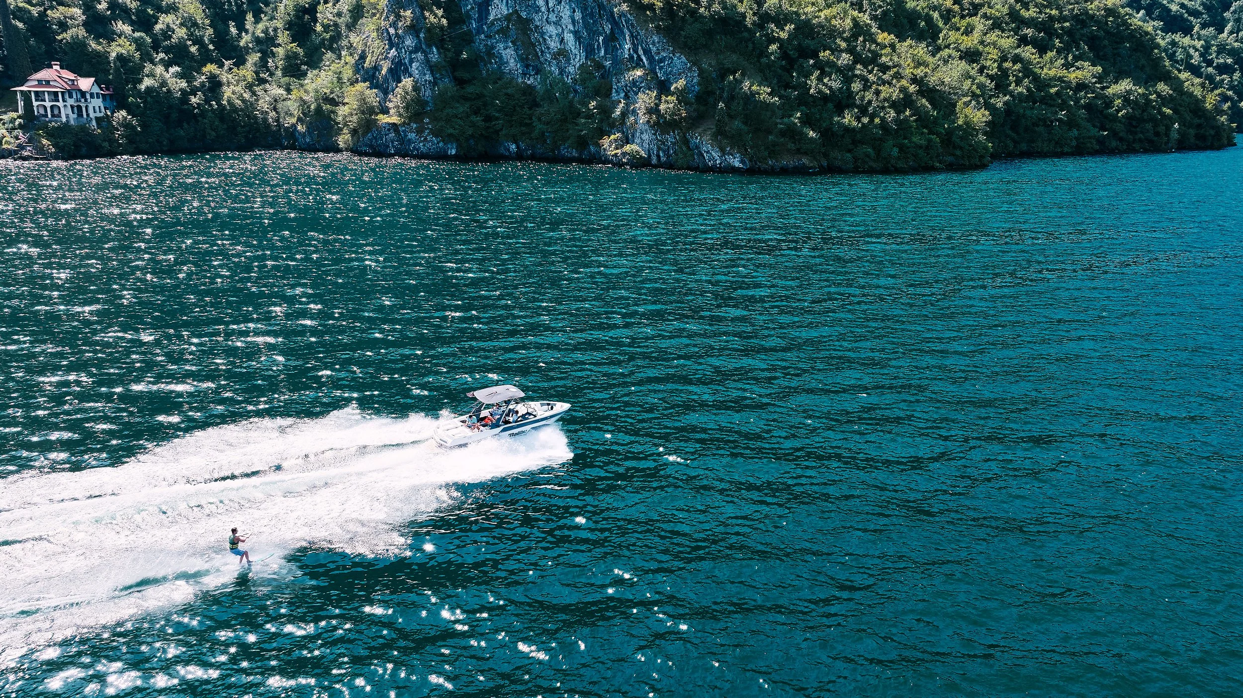 A person wakeboarding on a large body of water near a coastal area with green trees and steep cliffs; a motorboat is pulling the wakeboarder.