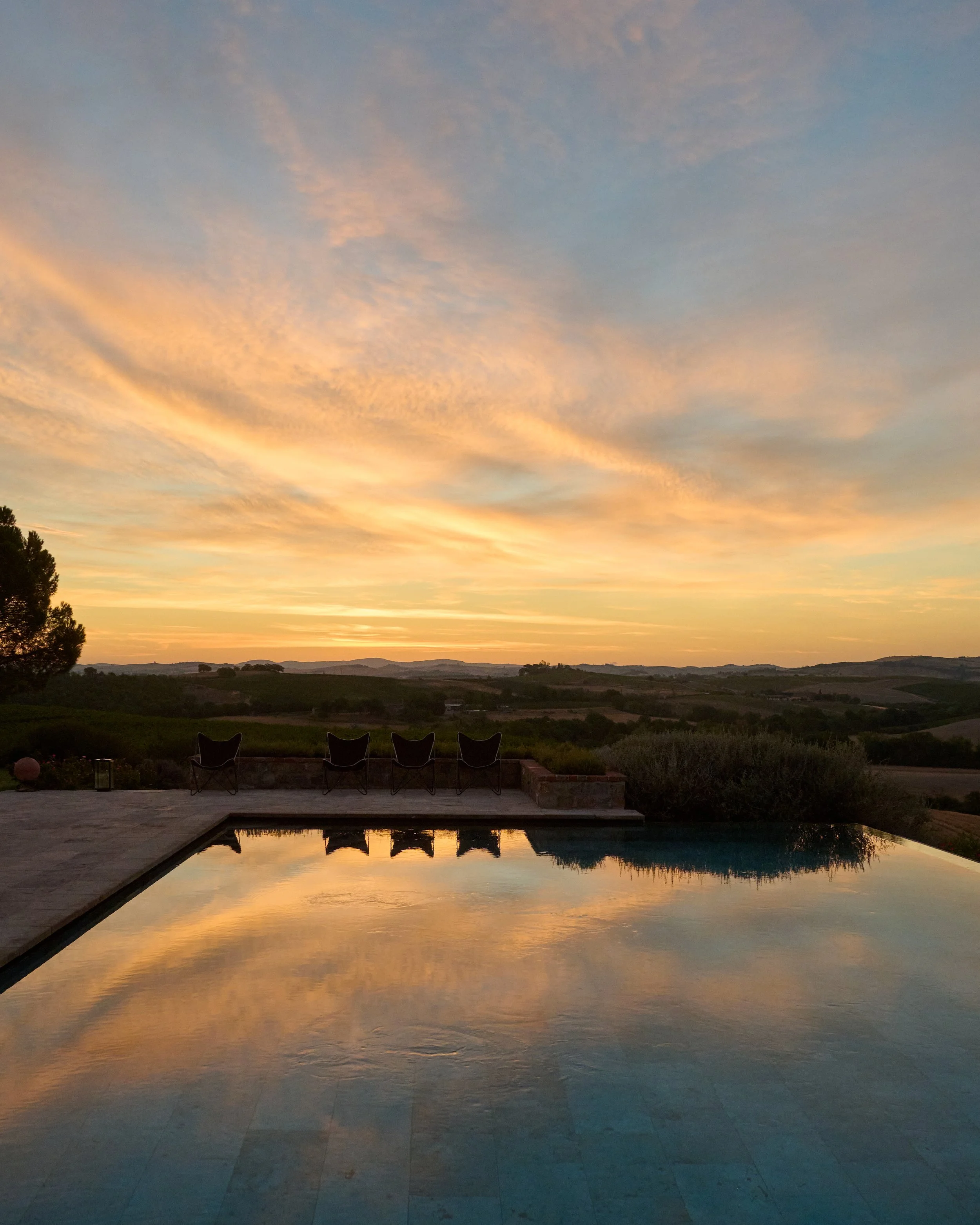 Sunset over a landscape with hills, reflected in an infinity pool with four chairs on the patio.