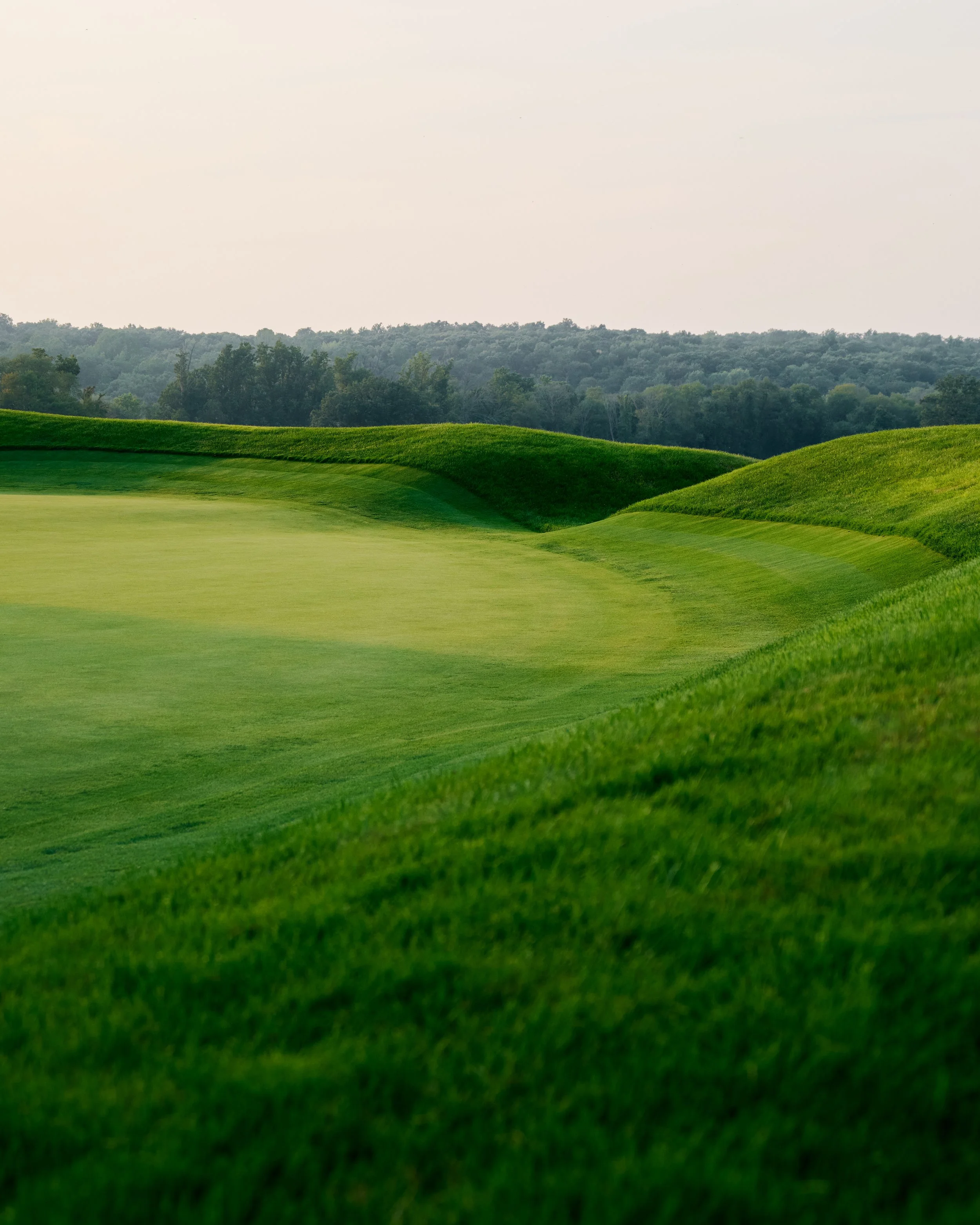 A lush green golf course with rolling hills and a distant tree line under a partly cloudy sky.