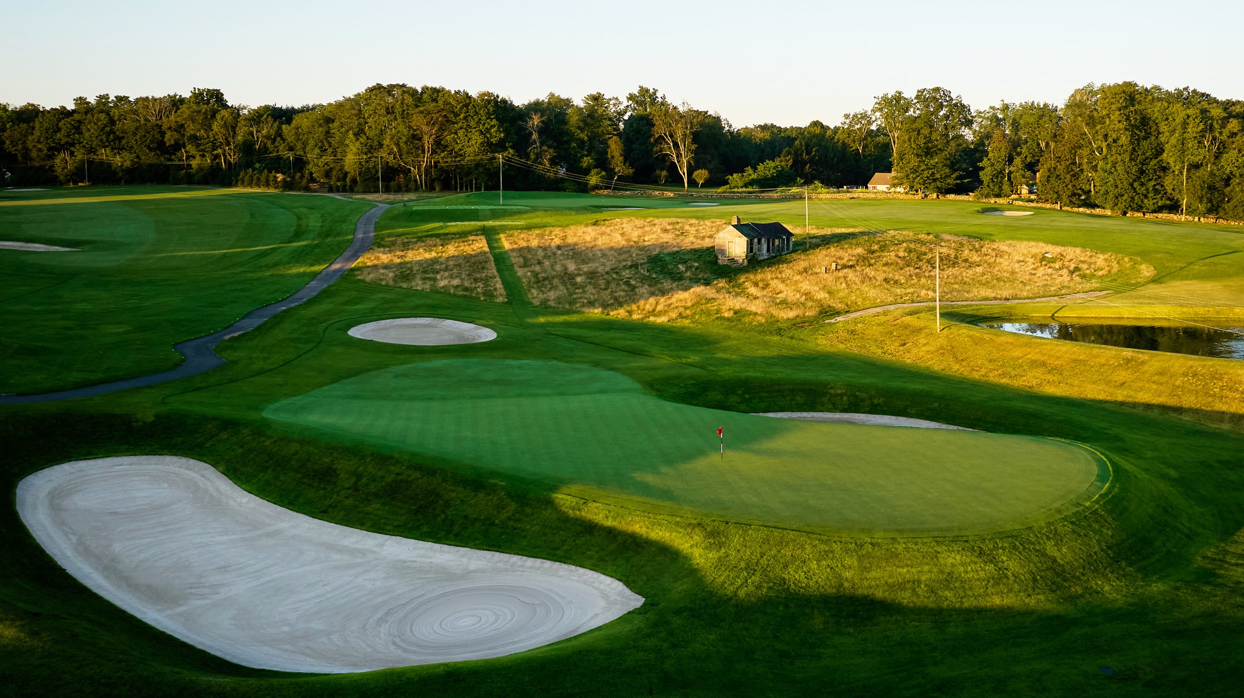 A scenic golf course with green fairways, sand bunkers, a putting green with a flag, a small pond, and a small building surrounded by trees in the background, illuminated by late afternoon sunlight.
