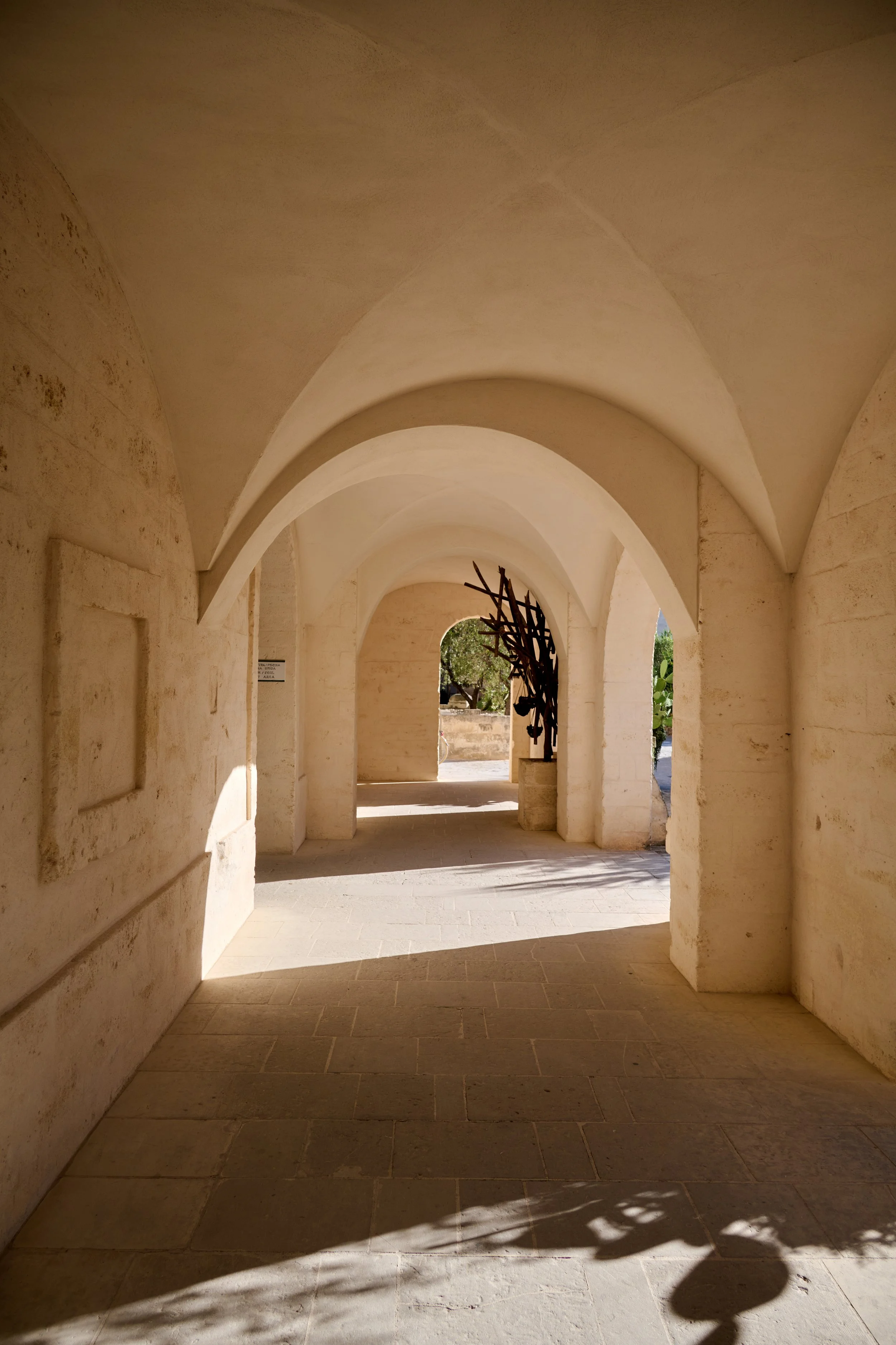 A stone archway with beige walls and ceiling, leading to an outdoor area with trees and a metal sculpture resembling a tree, casting shadows on the ground.