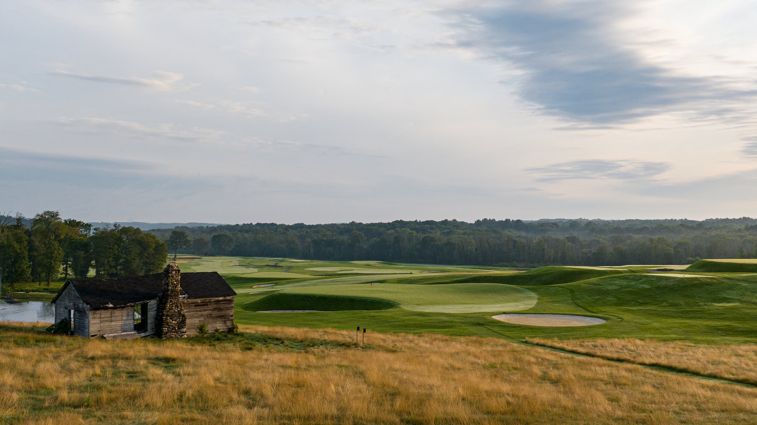 A scenic view of a golf course with green fairways, sand traps, and a small, weathered wooden house with a stone chimney in the foreground, surrounded by tall grass and trees under a partly cloudy sky.