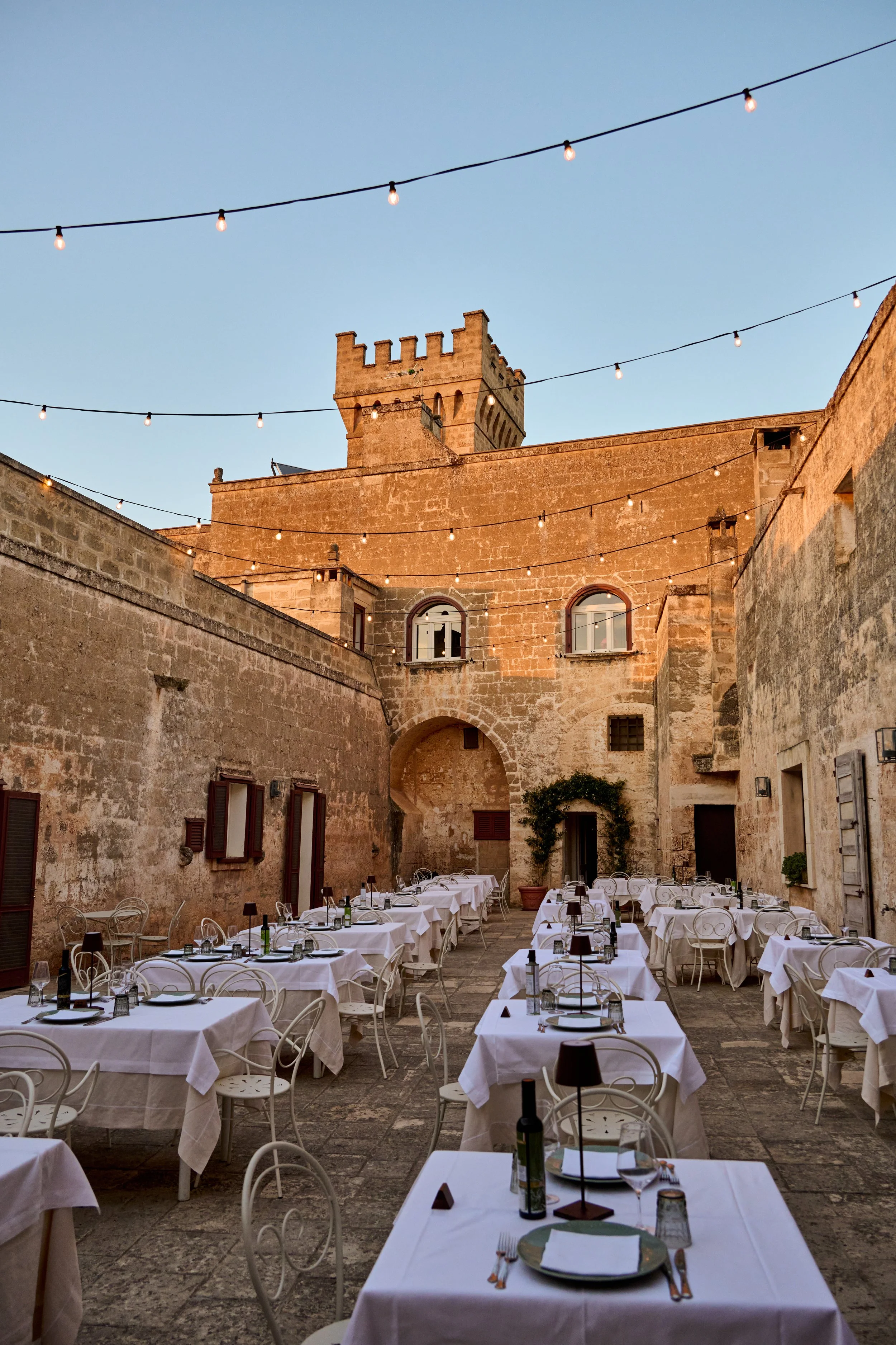 Outdoor restaurant with white tablecloths, black lamps, and wine bottles, enclosed by stone walls with a castle-like tower and string lights overhead during sunset.