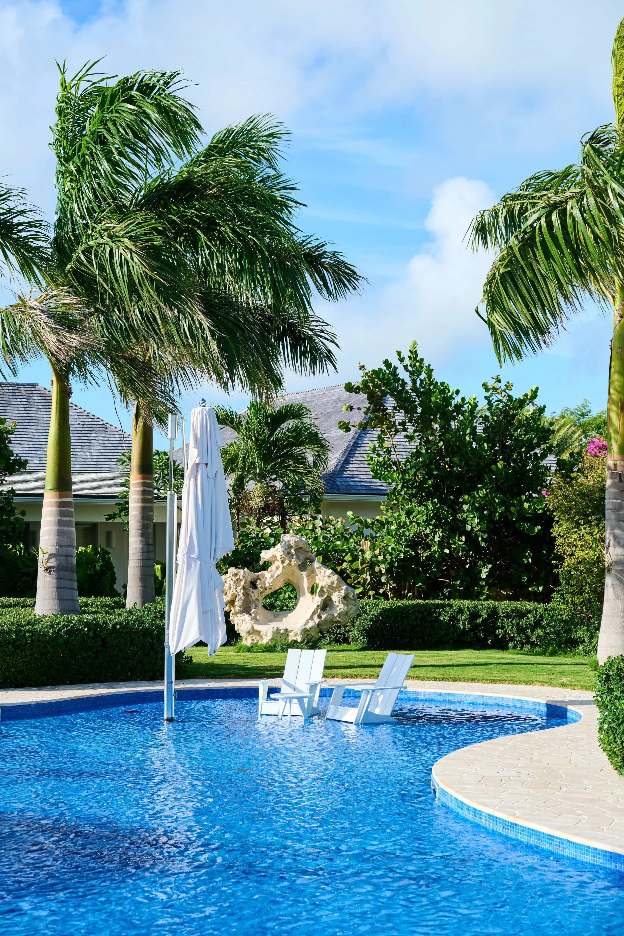 A swimming pool with two white lounge chairs and a closed white umbrella, surrounded by palm trees, lush green bushes, and a decorative stone sculpture, under a partly cloudy blue sky.