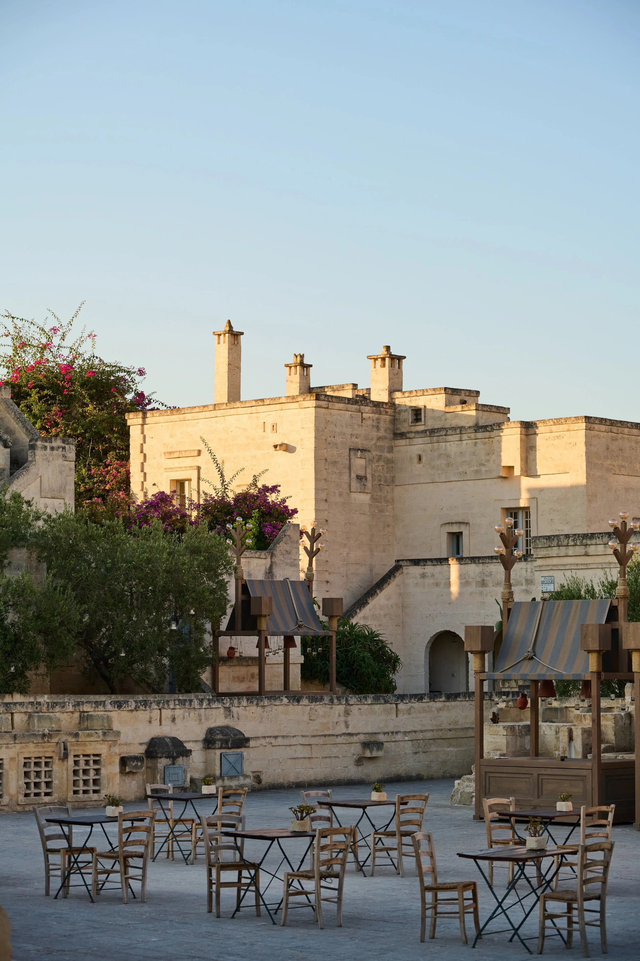 Outdoor cafe with tables and chairs near historic stone buildings with chimneys, trees, and colorful flowering bushes under a clear sky.