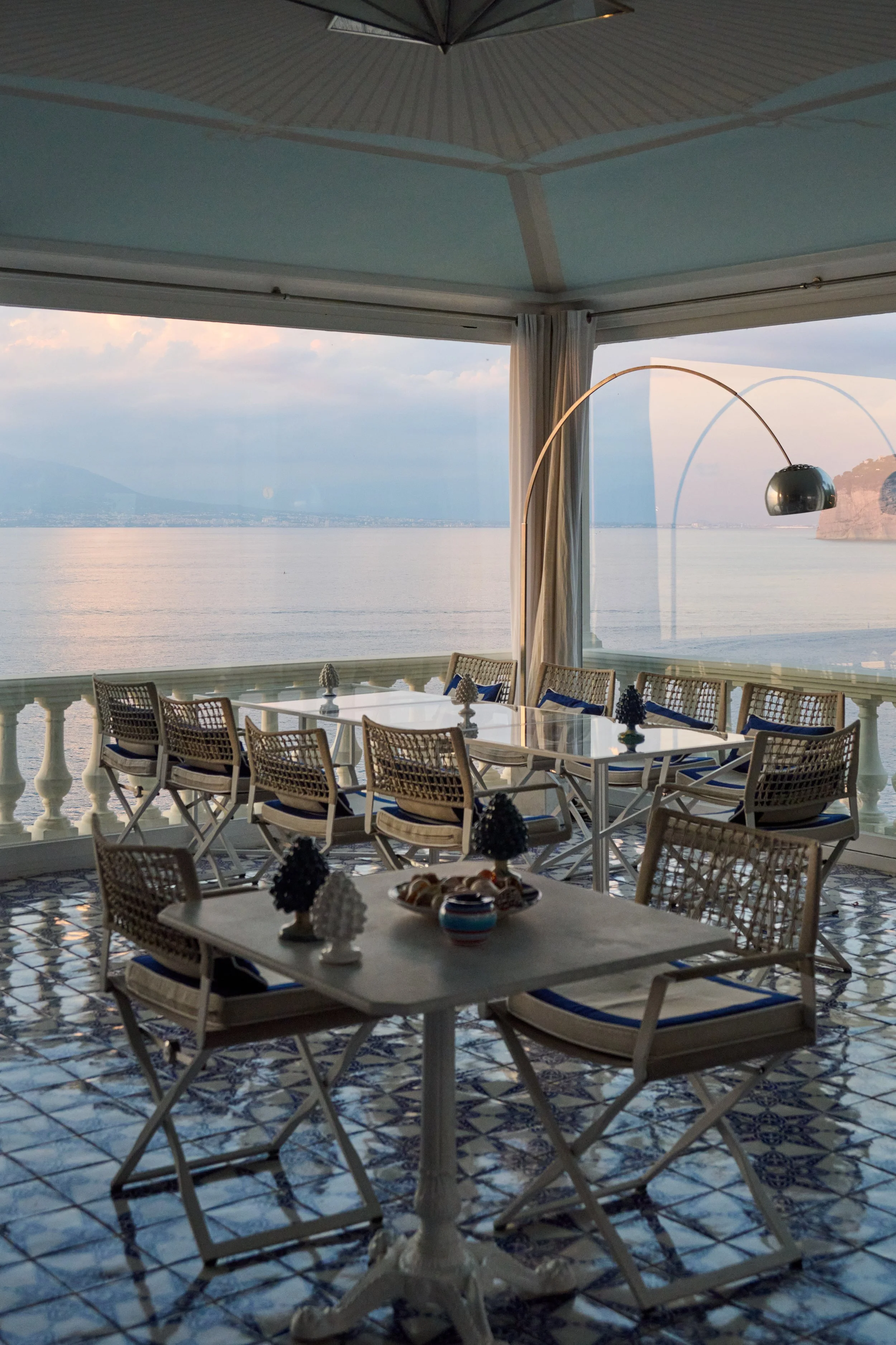 A bright, airy dining area with a view of the ocean. The room features a white marble table with chairs, decorative objects, and a large window or glass wall overlooking the water and distant mountains.