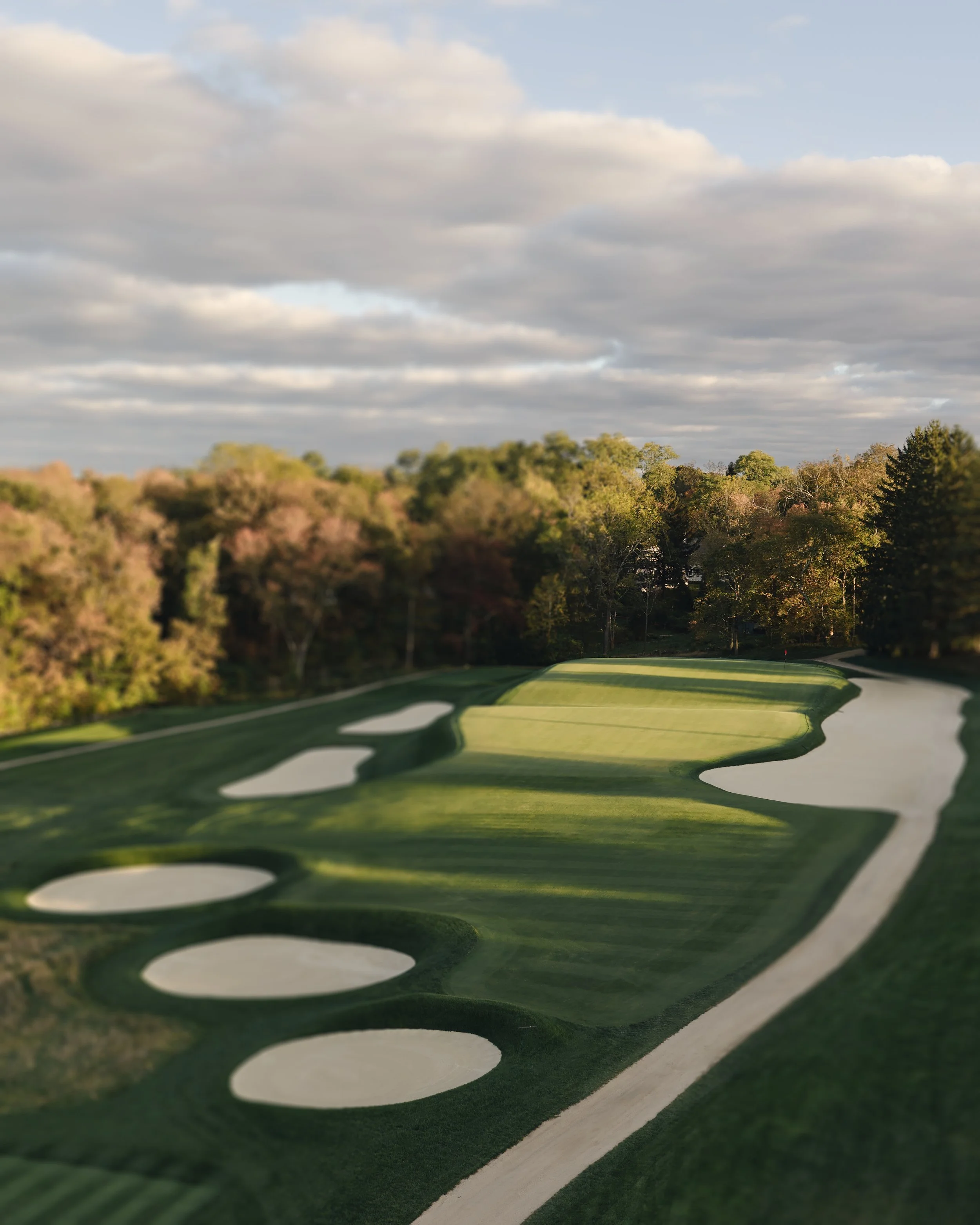A golf course with sand traps, green fairways, and a winding path, surrounded by trees under a cloudy sky.