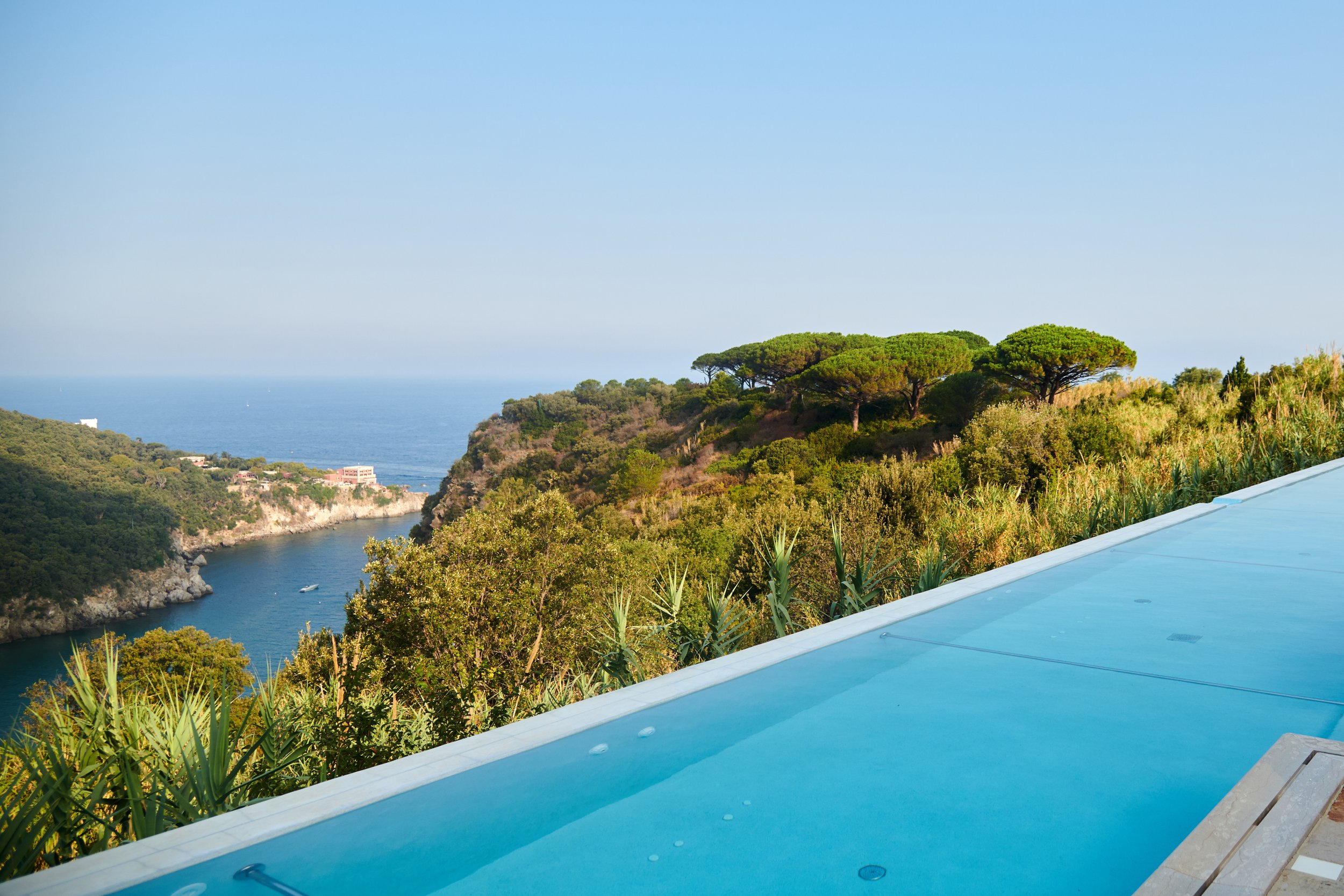 Infinity pool overlooking a lush green hillside with trees, a body of water, and the sea in the background under a clear blue sky.