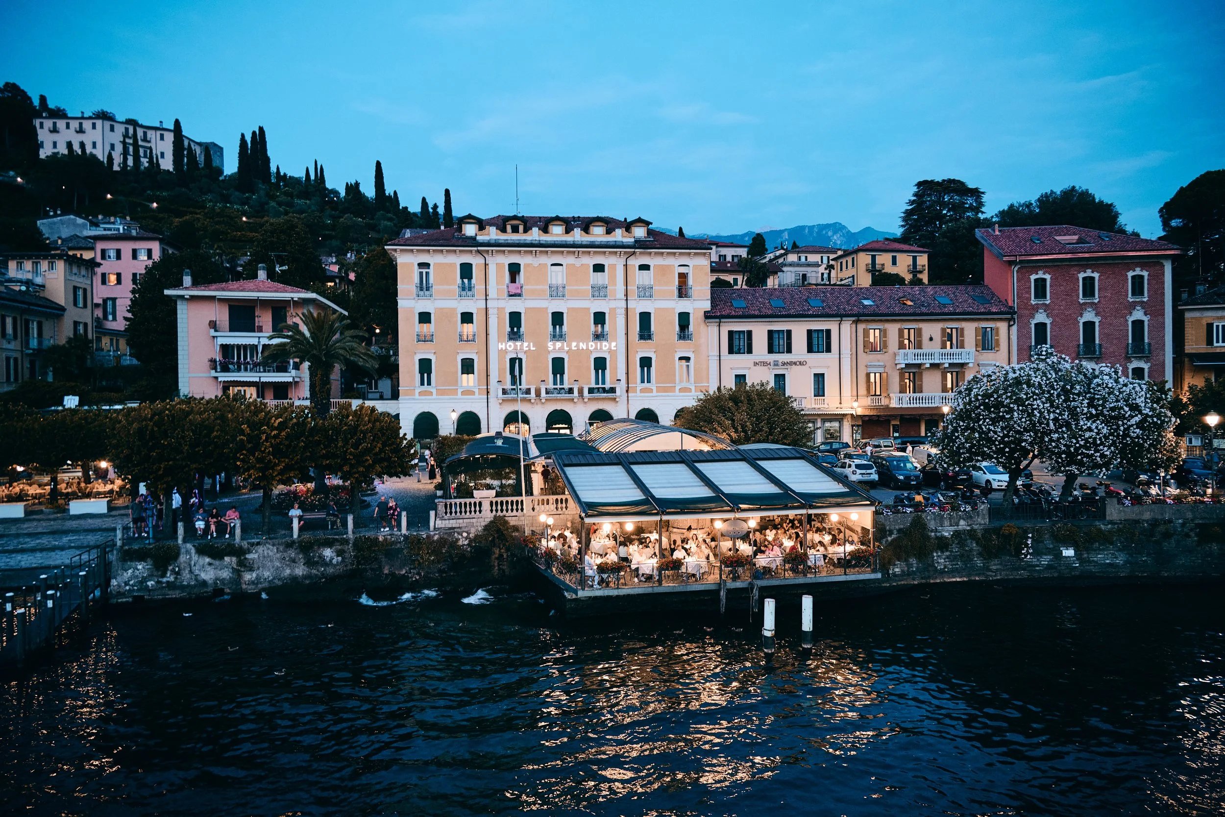 A waterfront terrace filled with diners at an outdoor restaurant during dusk, with colorful buildings and a hillside in the background.