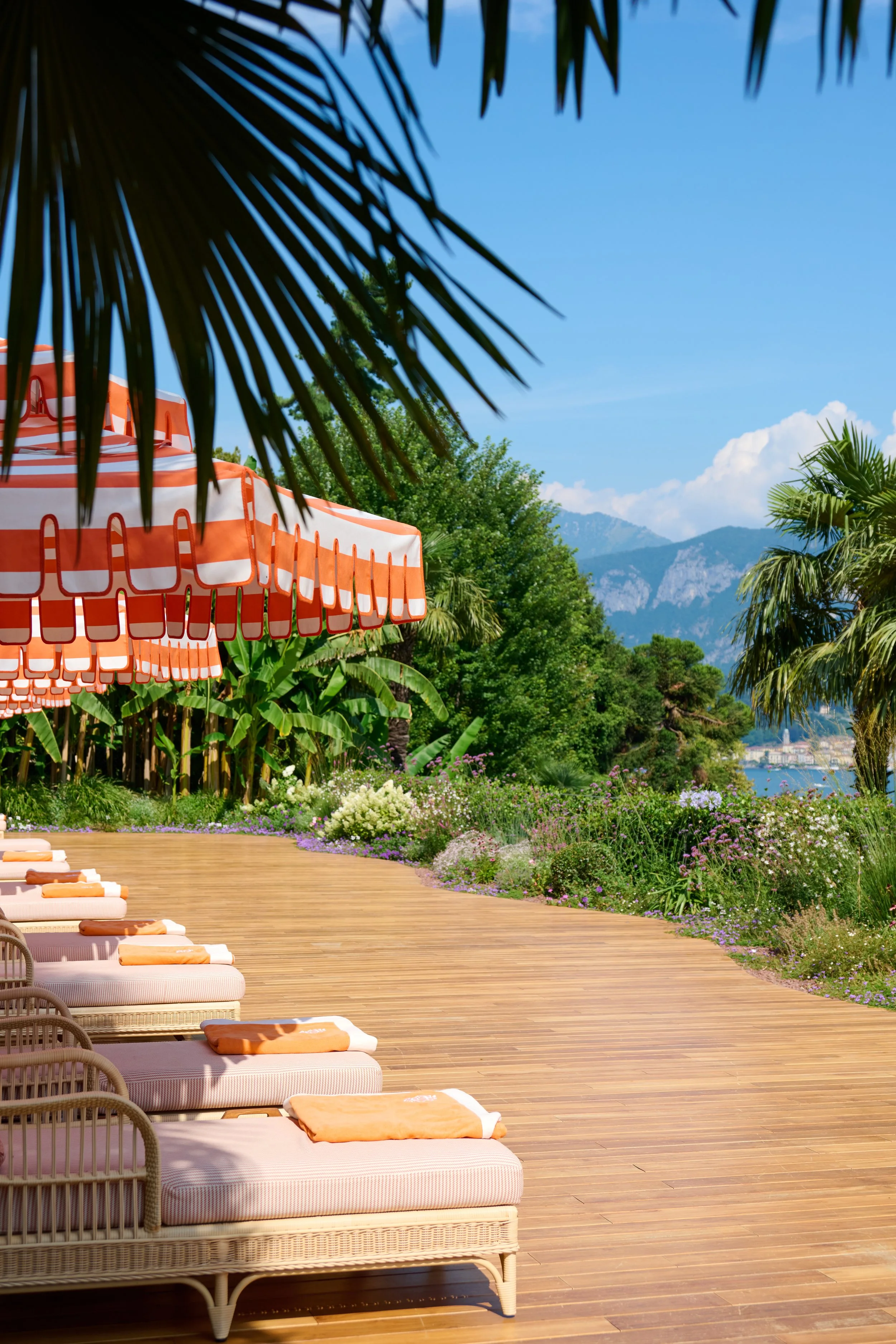 Outdoor lounge area with wicker daybeds and orange towels, large orange and white striped umbrellas, lush green foliage, flowering plants, and mountains in the background under a bright blue sky.