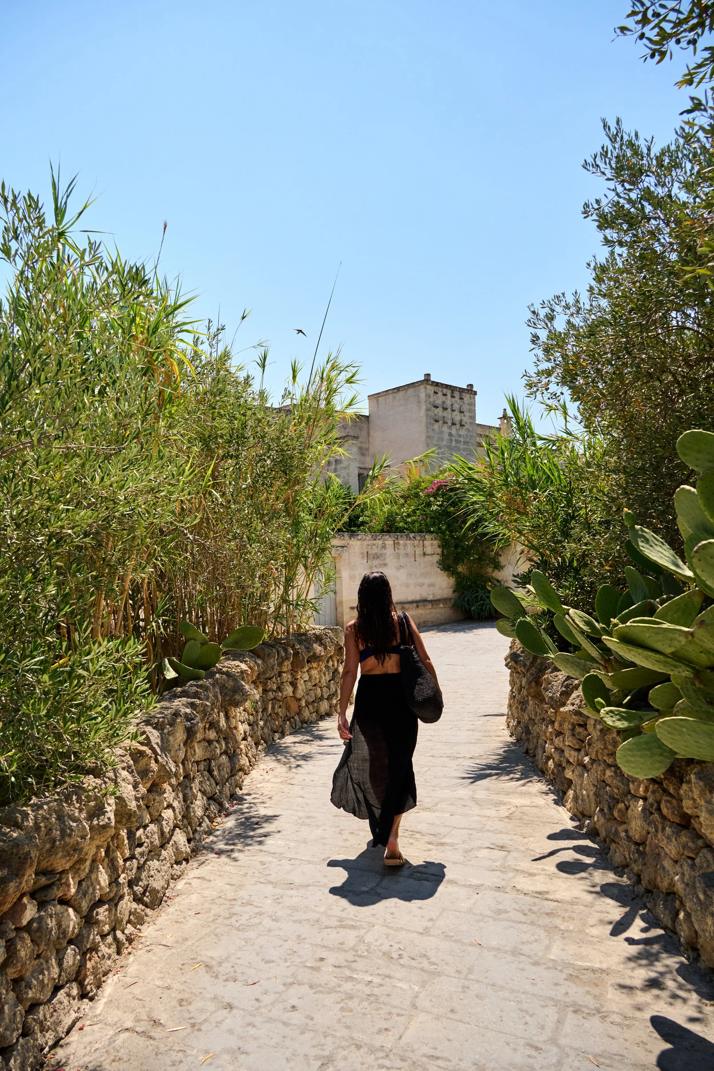 A woman in a black skirt and crop top walking on a stone path surrounded by green plants, with an old building and clear blue sky in the background.