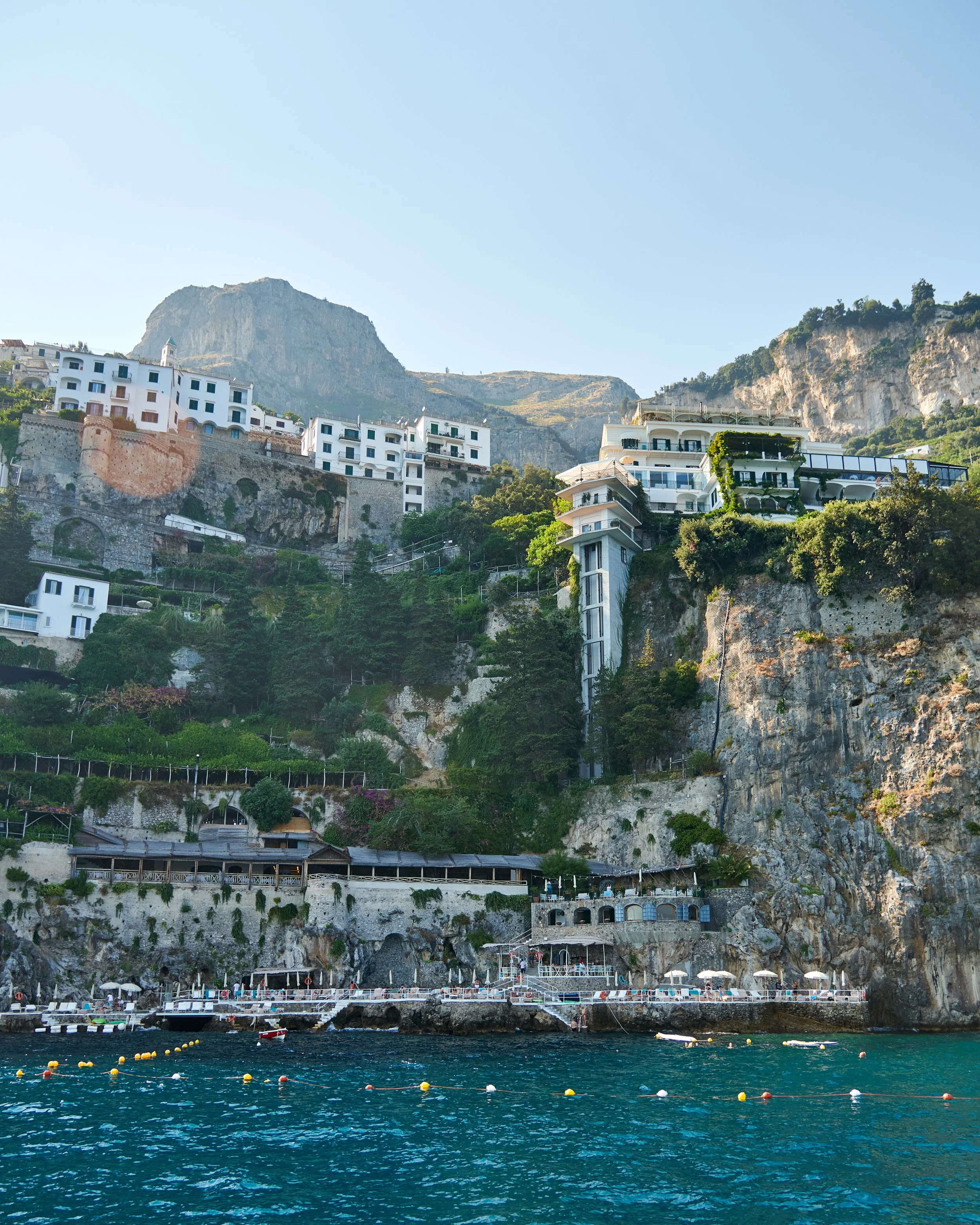 Coastal hillside with white buildings, greenery, and rocky cliffs overlooking the sea.