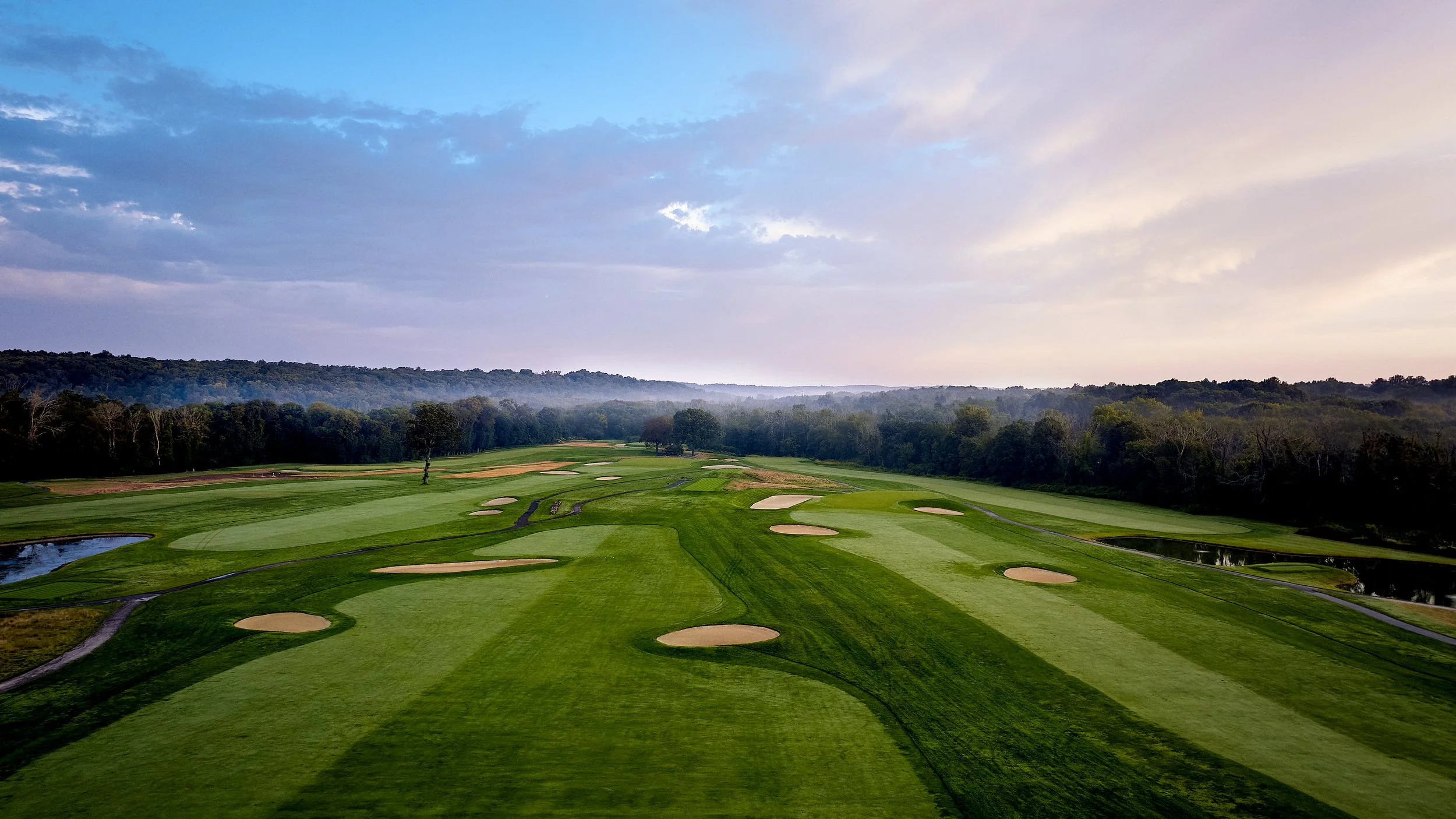 A golf course under a partly cloudy sky with bunkers, water hazards, and a well-maintained fairway, surrounded by trees.