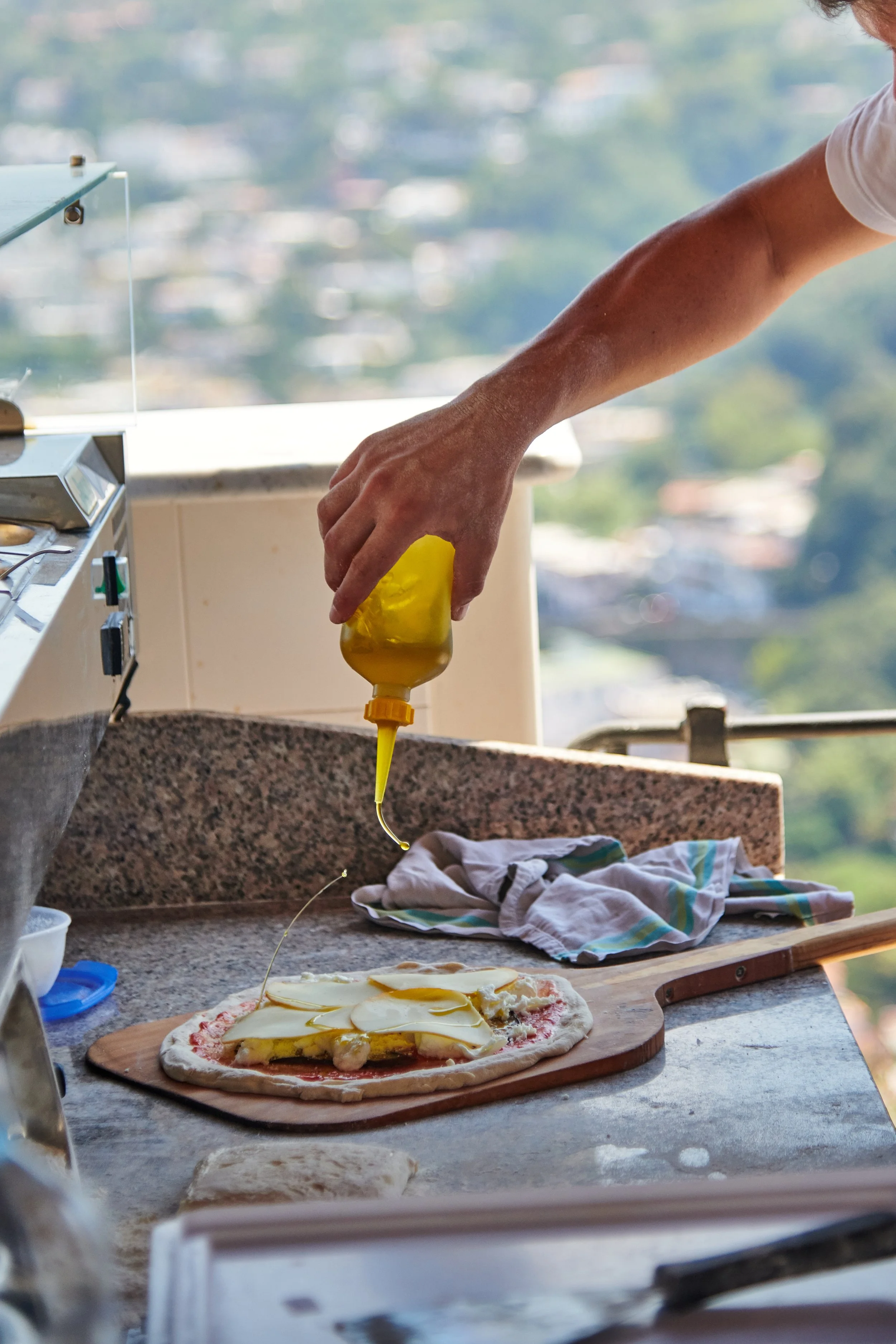 Person pouring yellow sauce or oil on a pizza crust topped with cheese and other ingredients, on a wooden cutting board in a kitchen with a city view in the background.