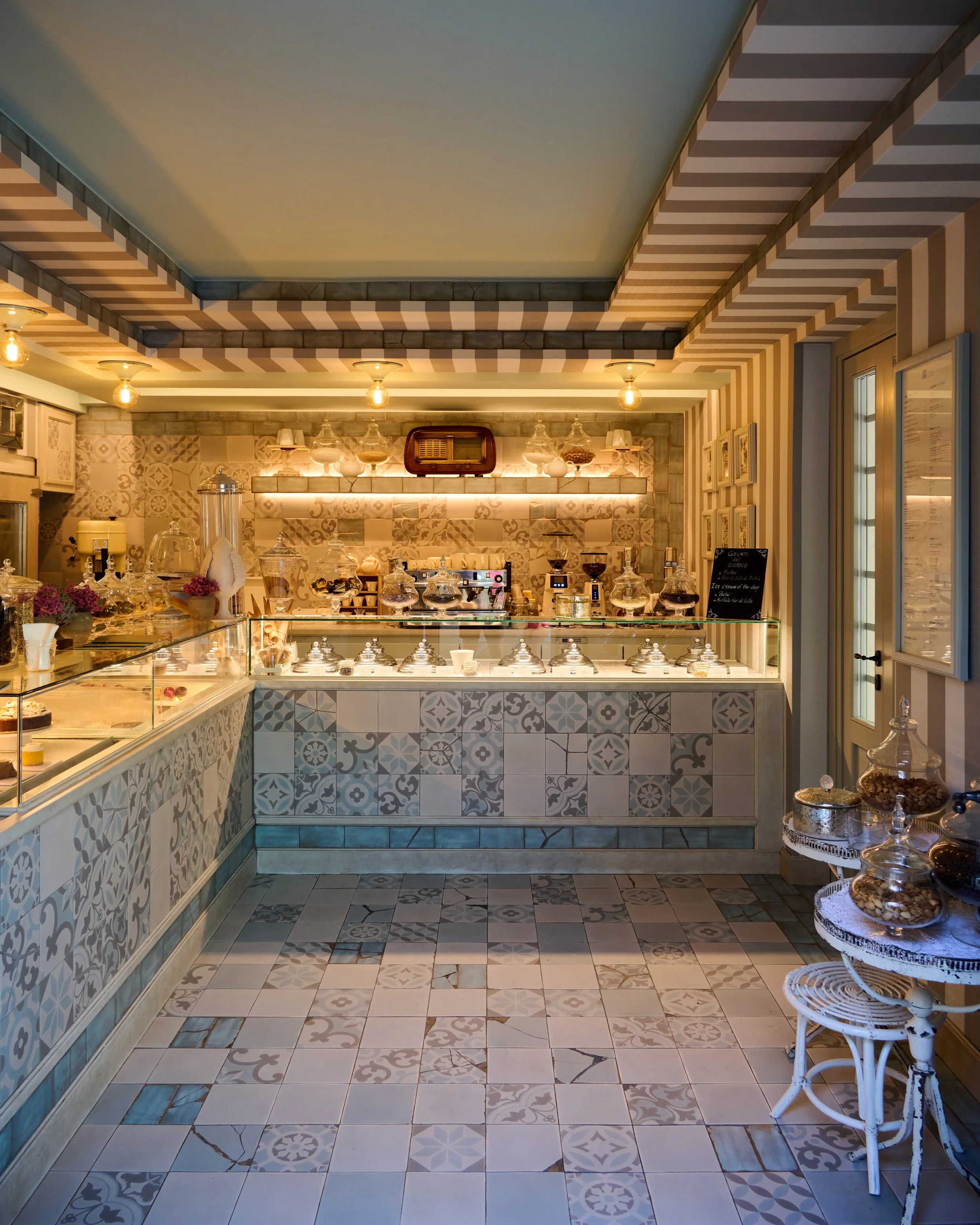 Interior of a bakery or pastry shop with a display case filled with cakes and desserts, decorated with jars and flowers, and a patterned tiled wall and floor.