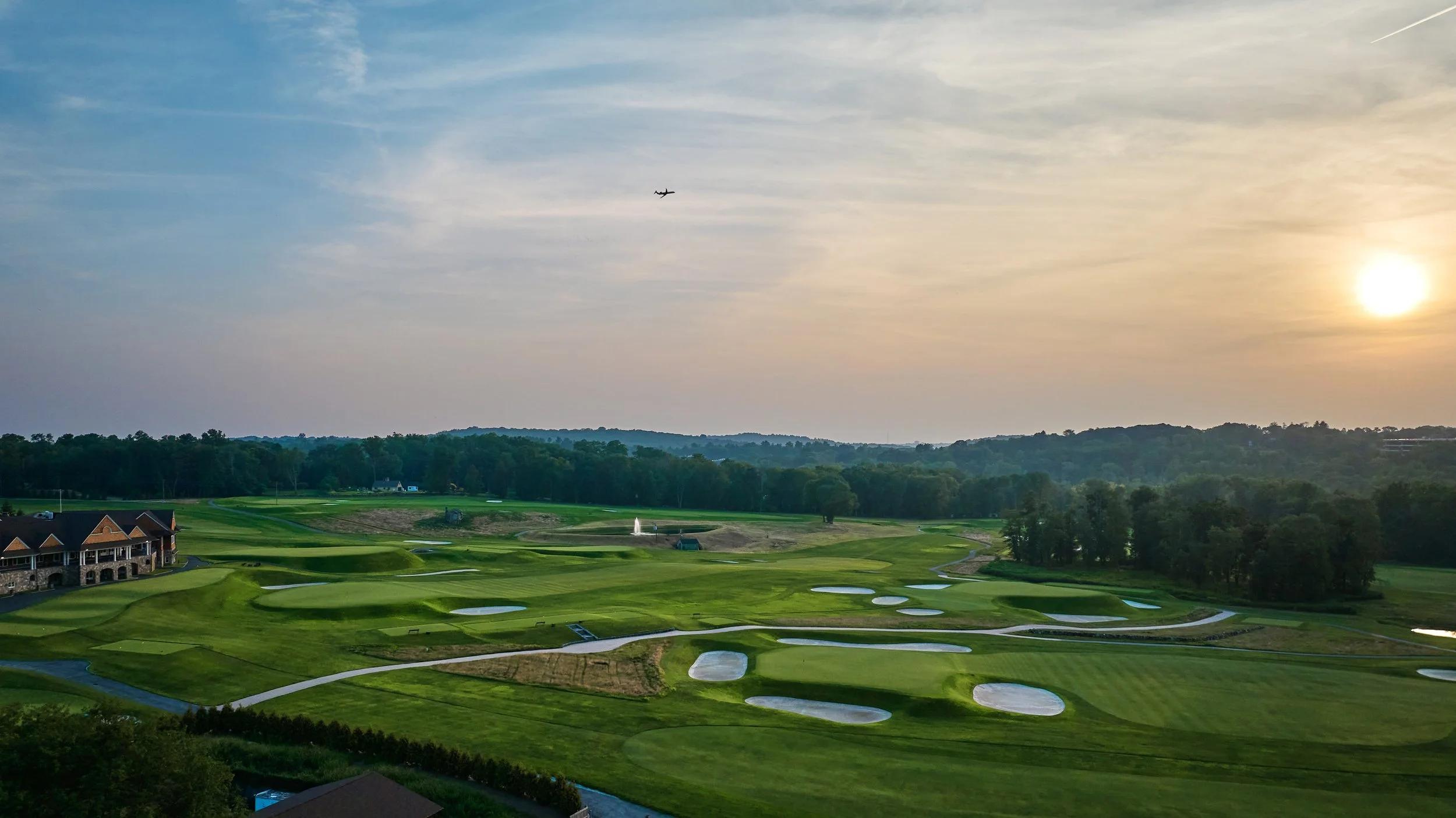 A scenic view of a golf course at sunset with sand traps, a water fountain, and a building in the distance, surrounded by green hills and trees.