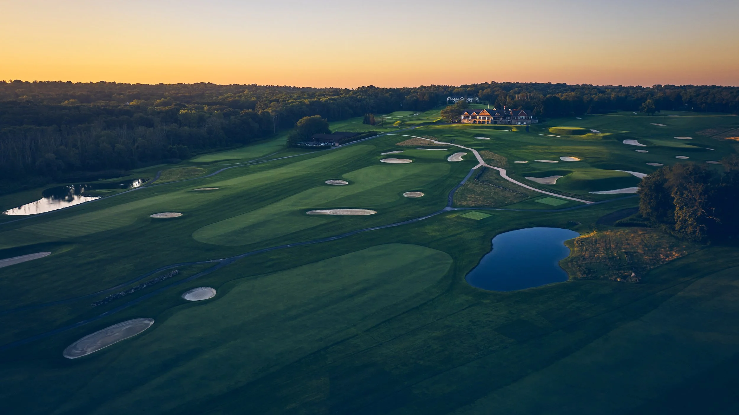 Aerial view of a golf course at sunset featuring green fairways, sand traps, water hazards, and a large clubhouse in the distance.