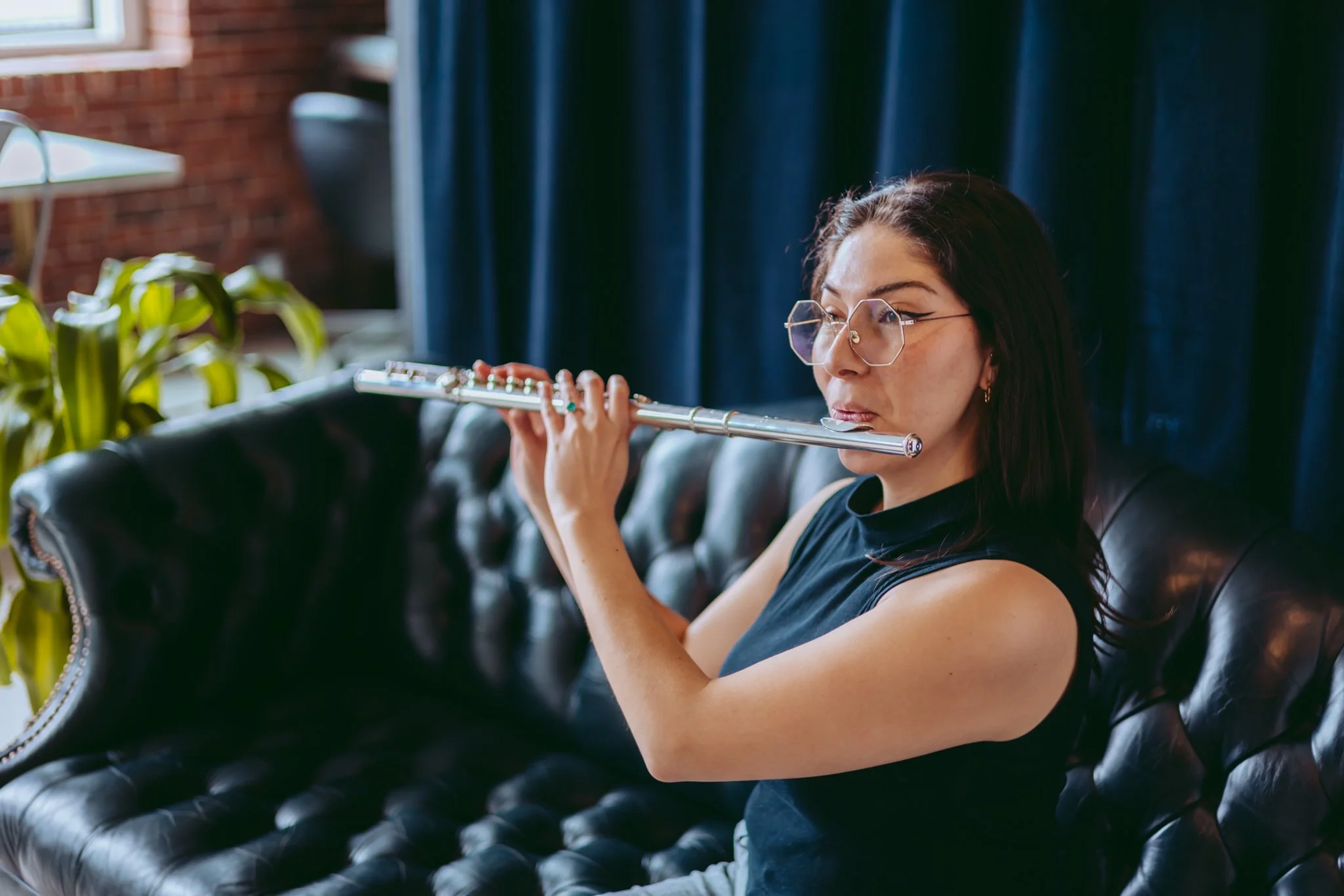 Woman with dark hair and glasses playing a silver flute while seated on a black leather couch in a room with brick walls and blue curtains, near a window with sunlight.
