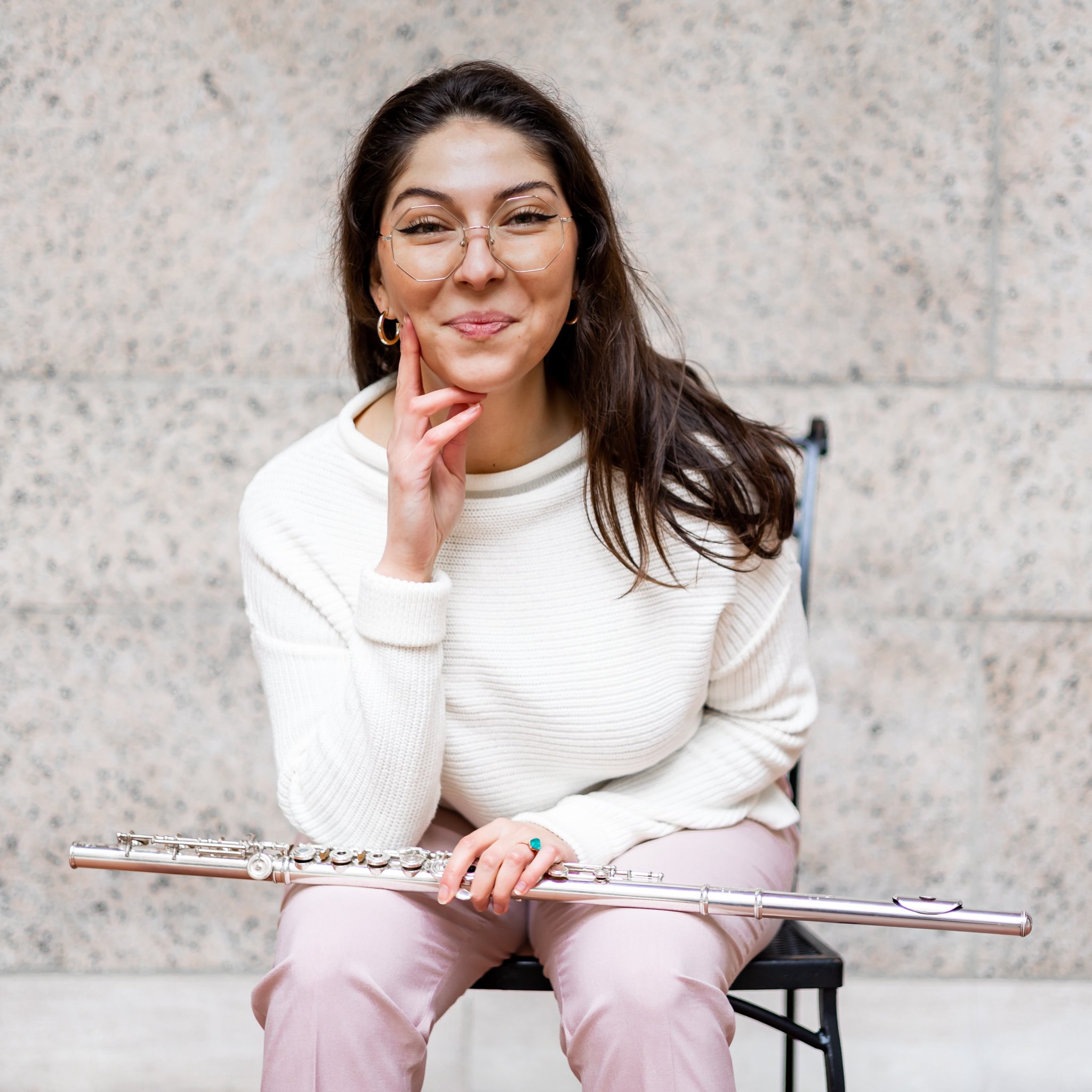 Woman with glasses smiling and touching her cheek, sitting on a chair holding a flute, wearing a white sweater and light pink pants, against a textured wall background.