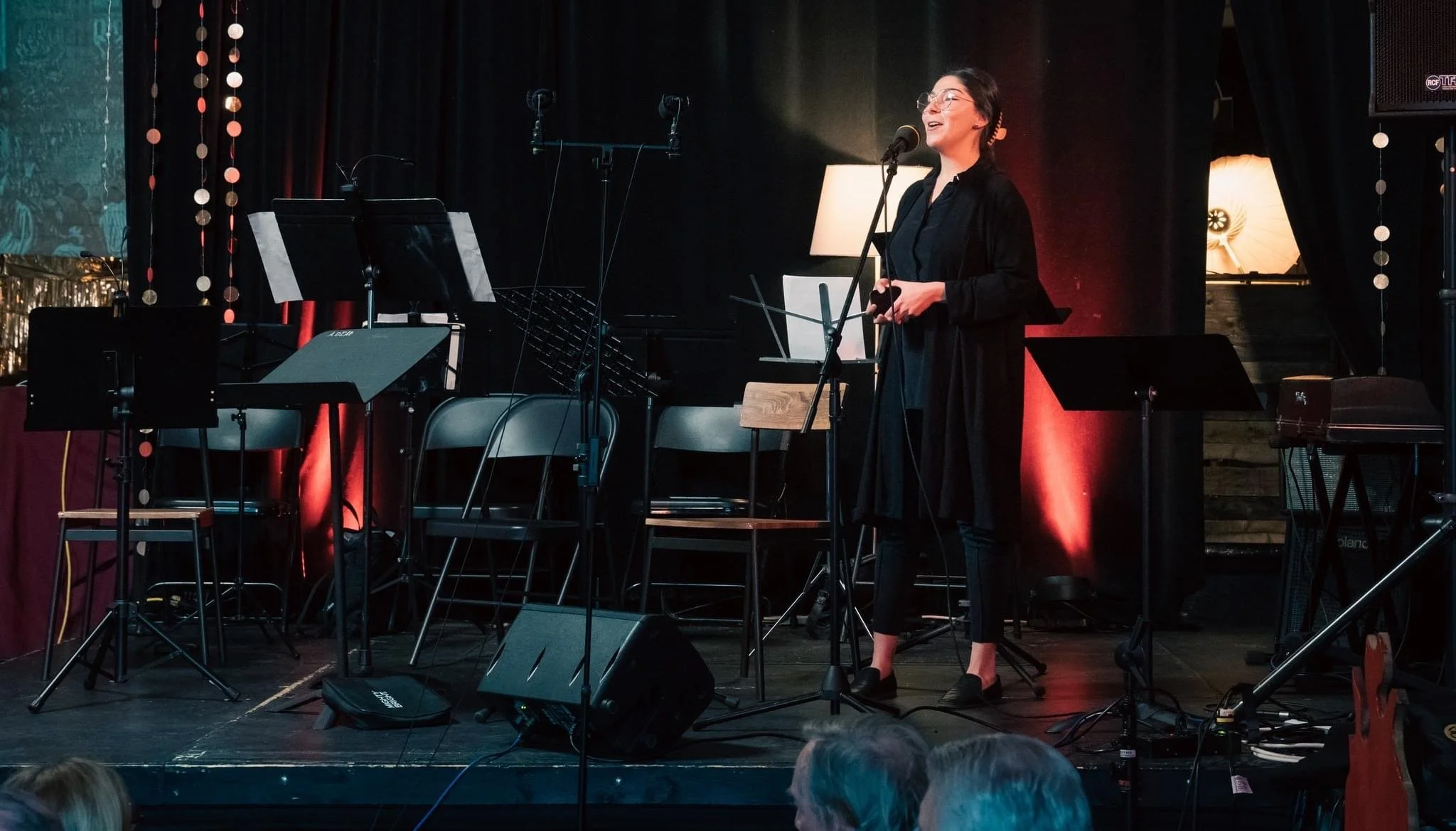 A woman in glasses and black clothing singing into a microphone on a stage decorated with red and gold hanging ornaments, surrounded by musical instruments and equipment.