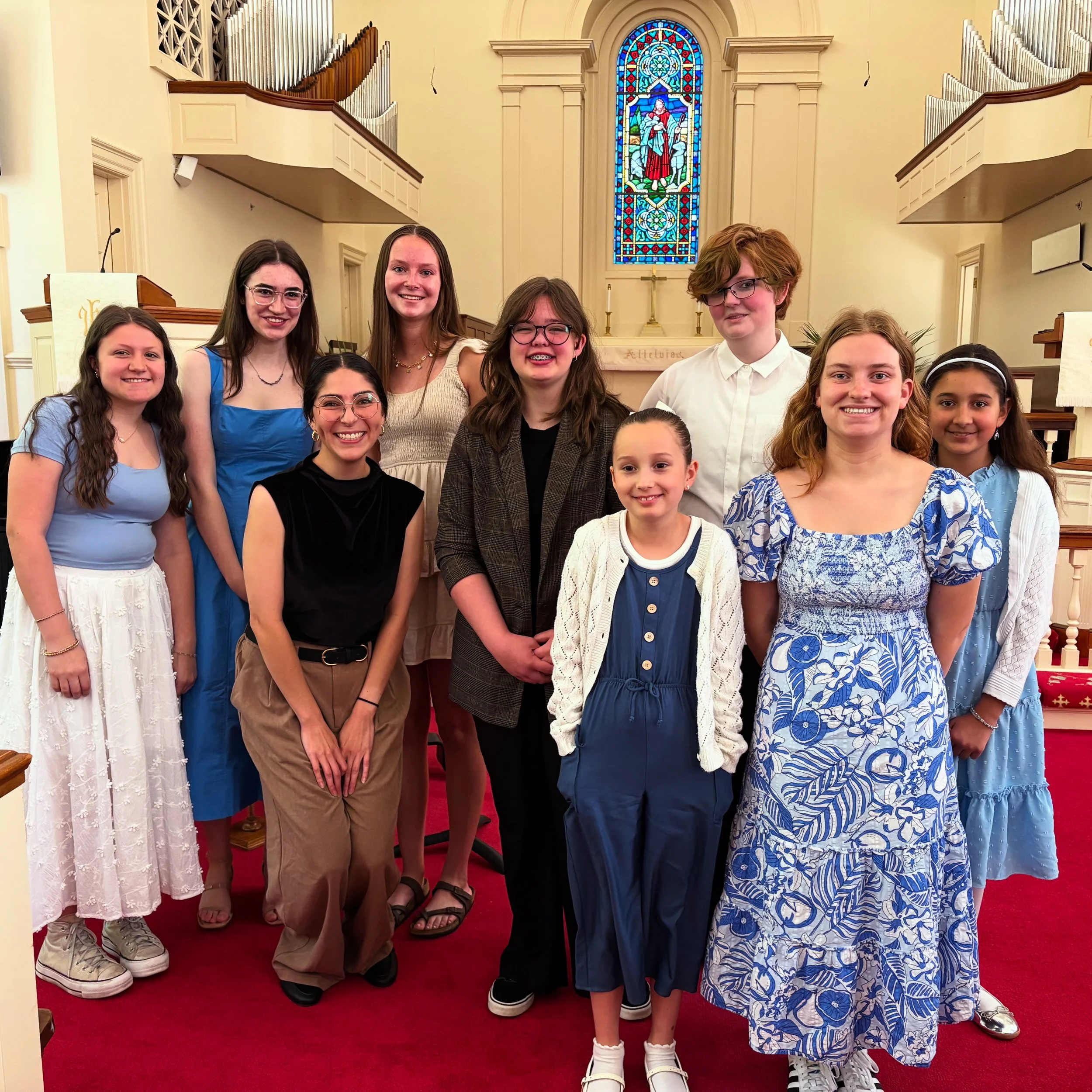 Group of children and teenagers standing inside a church with a colorful stained glass window and an altar in the background. The group is smiling and dressed in casual summer clothing. This picture was taken from the DiazFlute Spring Flute Recital.