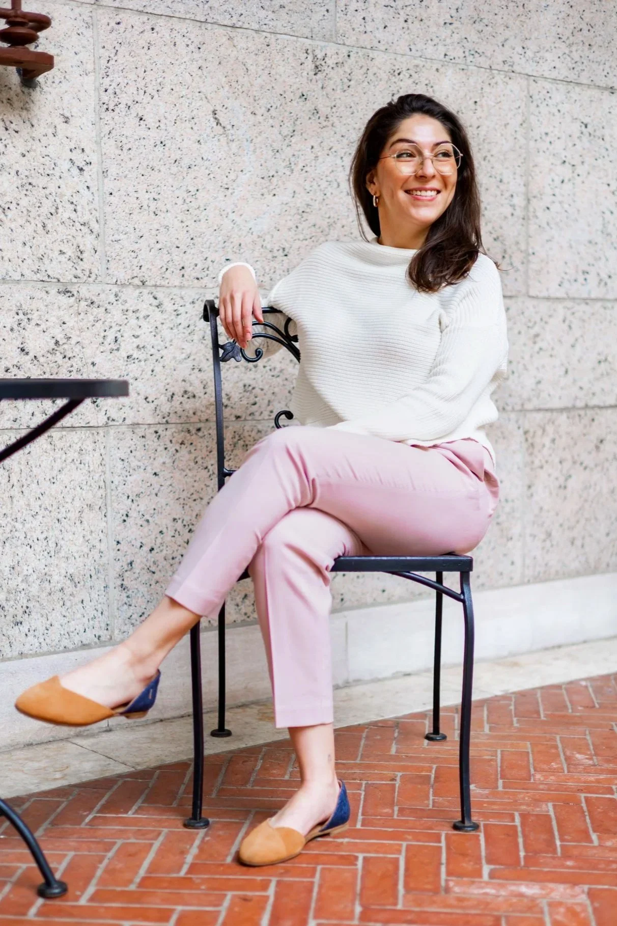 A woman with dark hair, glasses, and earrings is sitting on a black metal chair outdoors against a textured stone wall, smiling and looking to her right. She is wearing a white sweater, pink pants, and tan flats with dark blue accents.