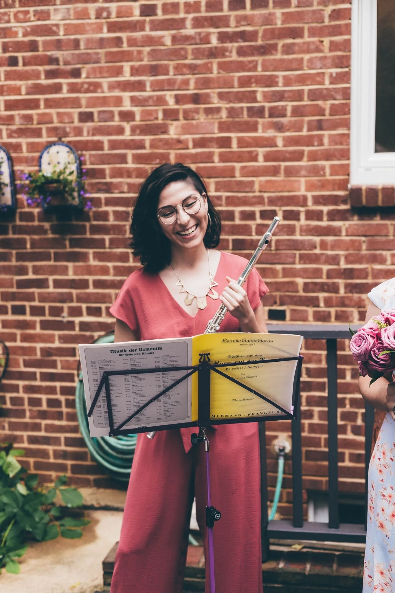 A woman playing a flute outdoors, standing in front of a brick wall, looking at sheet music on a music stand, smiling, wearing glasses and a red dress.