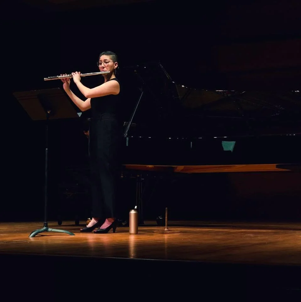 A woman playing the flute on a stage with a grand piano in the background.