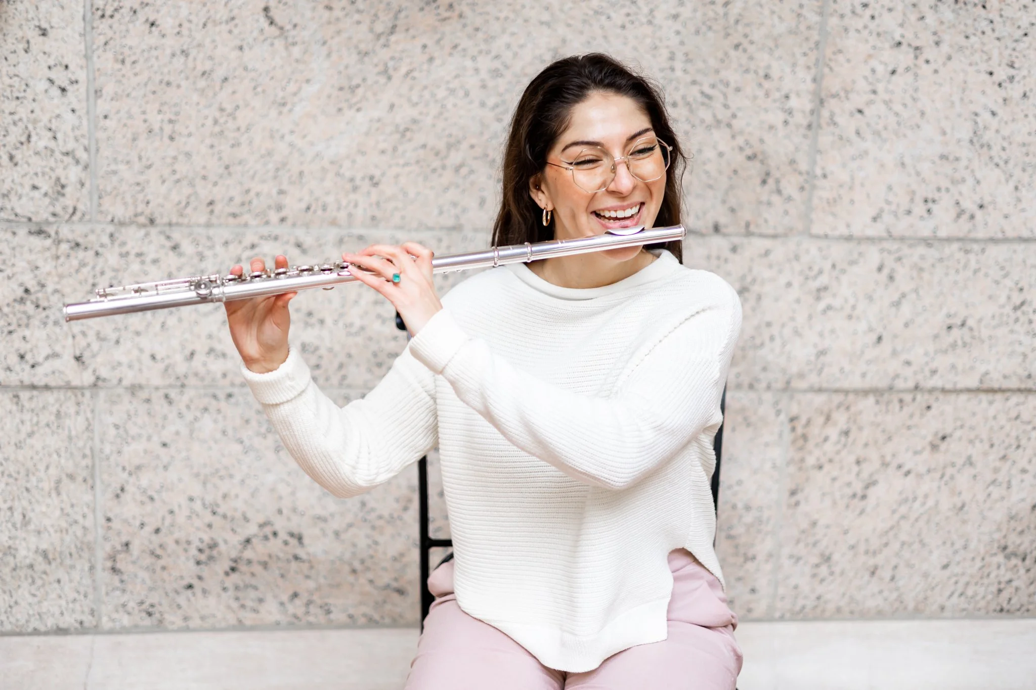 A woman with dark hair, glasses, and earrings playing a silver flute, smiling, seated against a speckled stone wall, wearing a white sweater and pink pants.