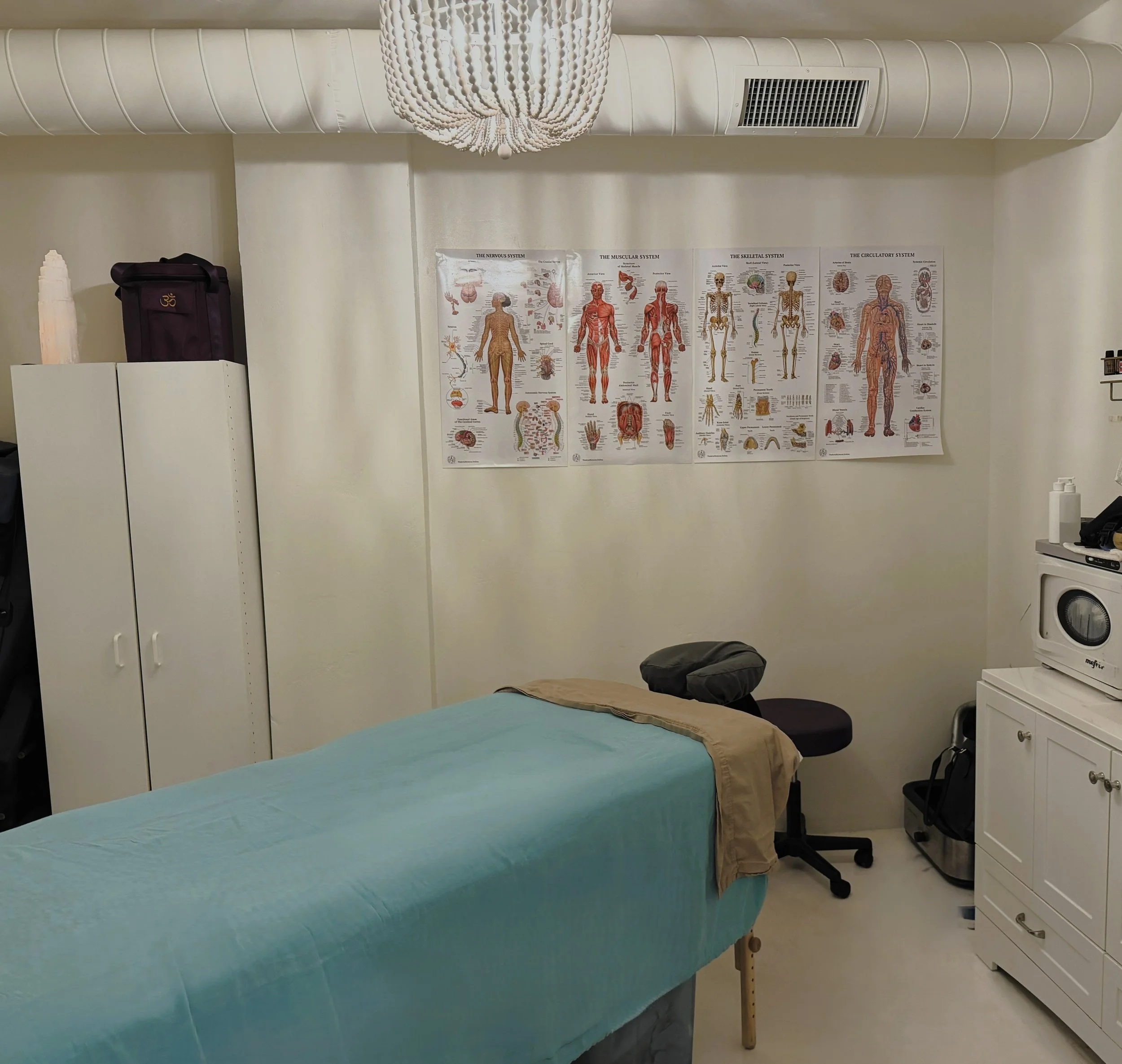 A room with a massage table covered in turquoise sheets, a white cabinet, posters of human body systems on the wall, and medical supplies.