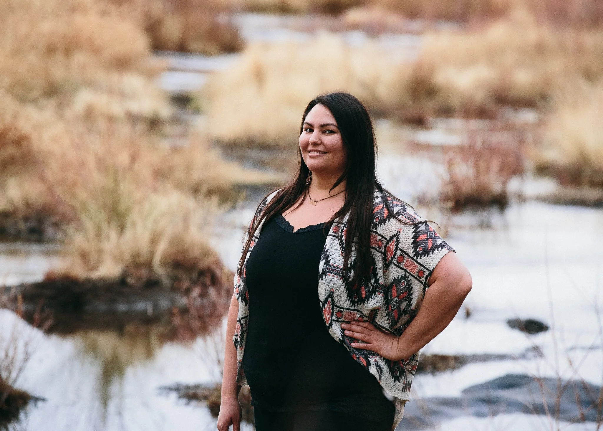 A woman with long dark hair smiling outdoors during winter, wearing a patterned cardigan over a navy top, with dry bushes and snow in the background.