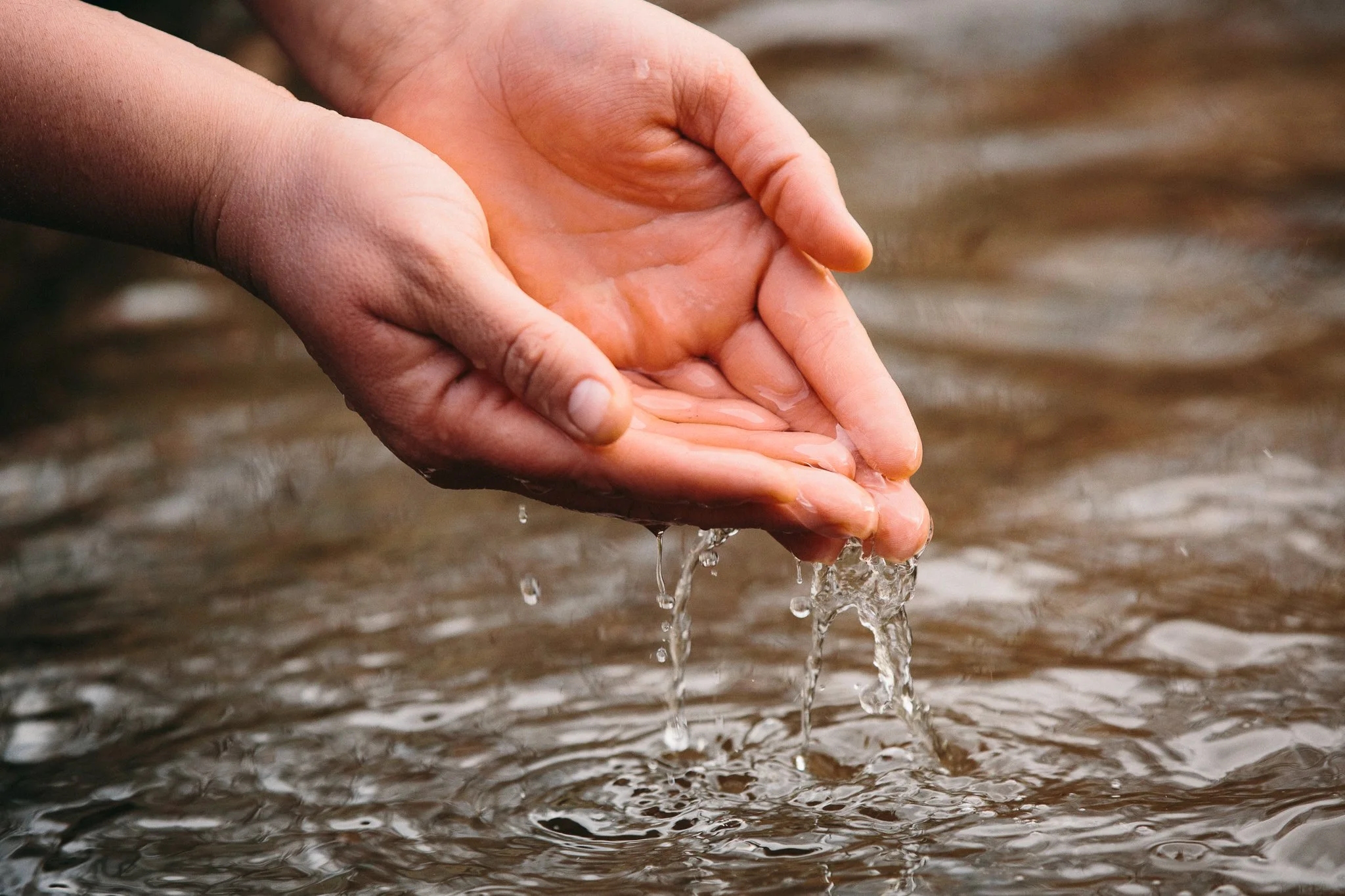Close-up of hands holding water, with water droplets dripping from the fingers over a body of water.