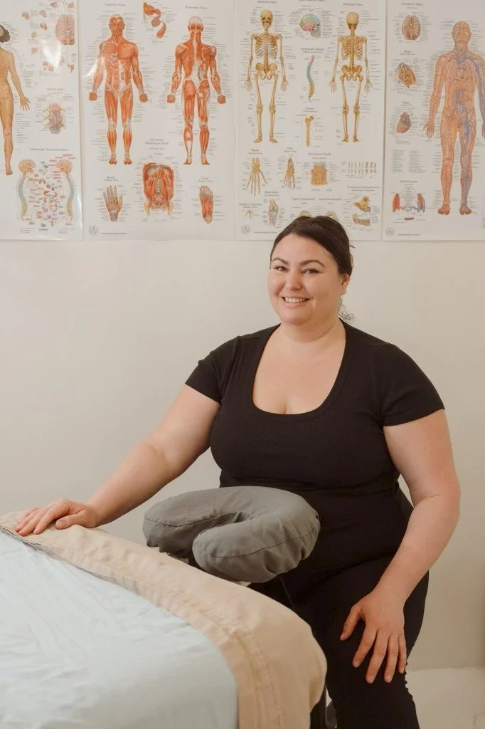 A woman smiling at a massage table with a pillow, in a room with anatomical posters on the wall.