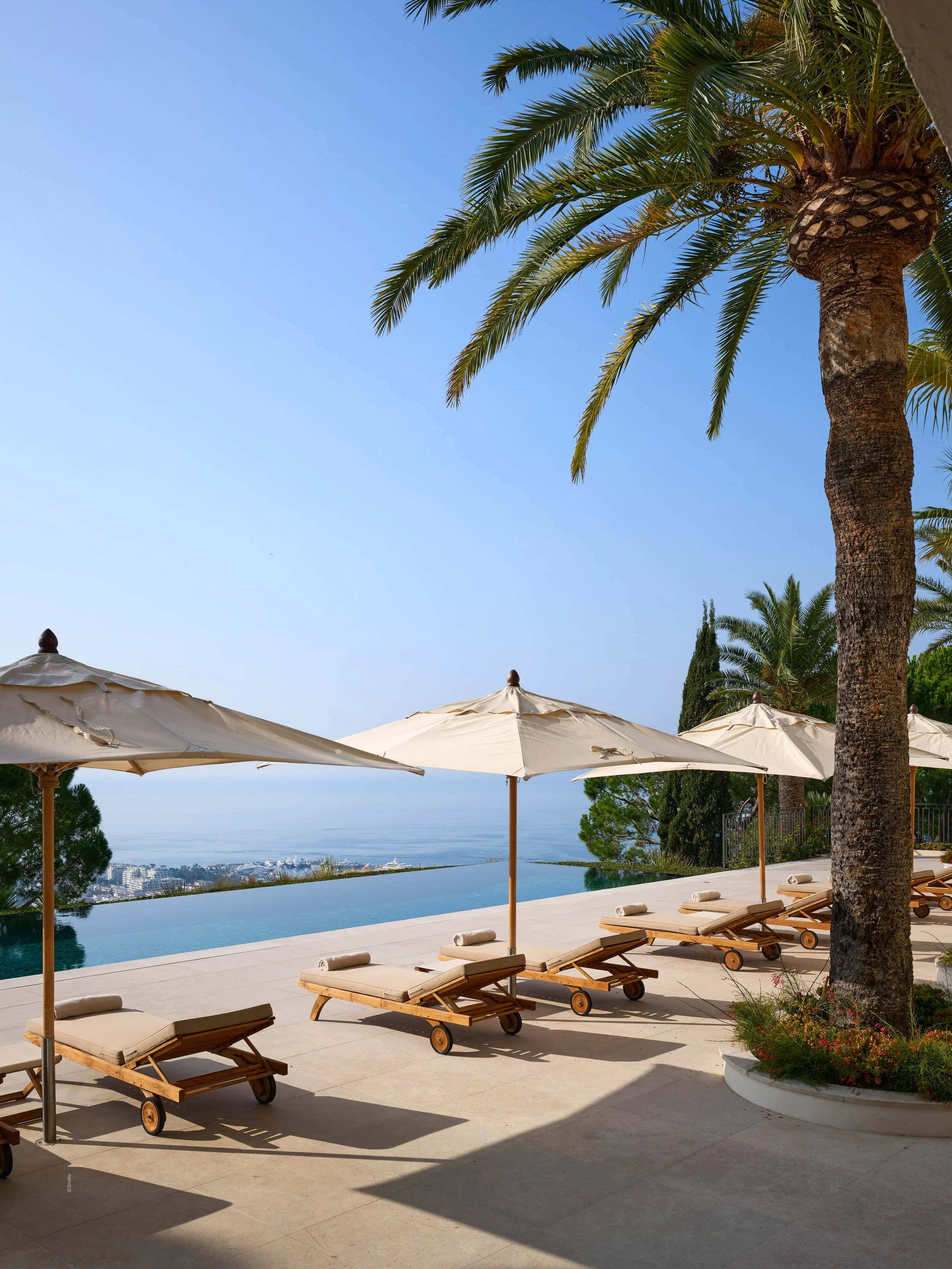 Chaises longues en bois avec coussins et parasols crème au bord de la piscine avec vue sur la mer et des palmiers.