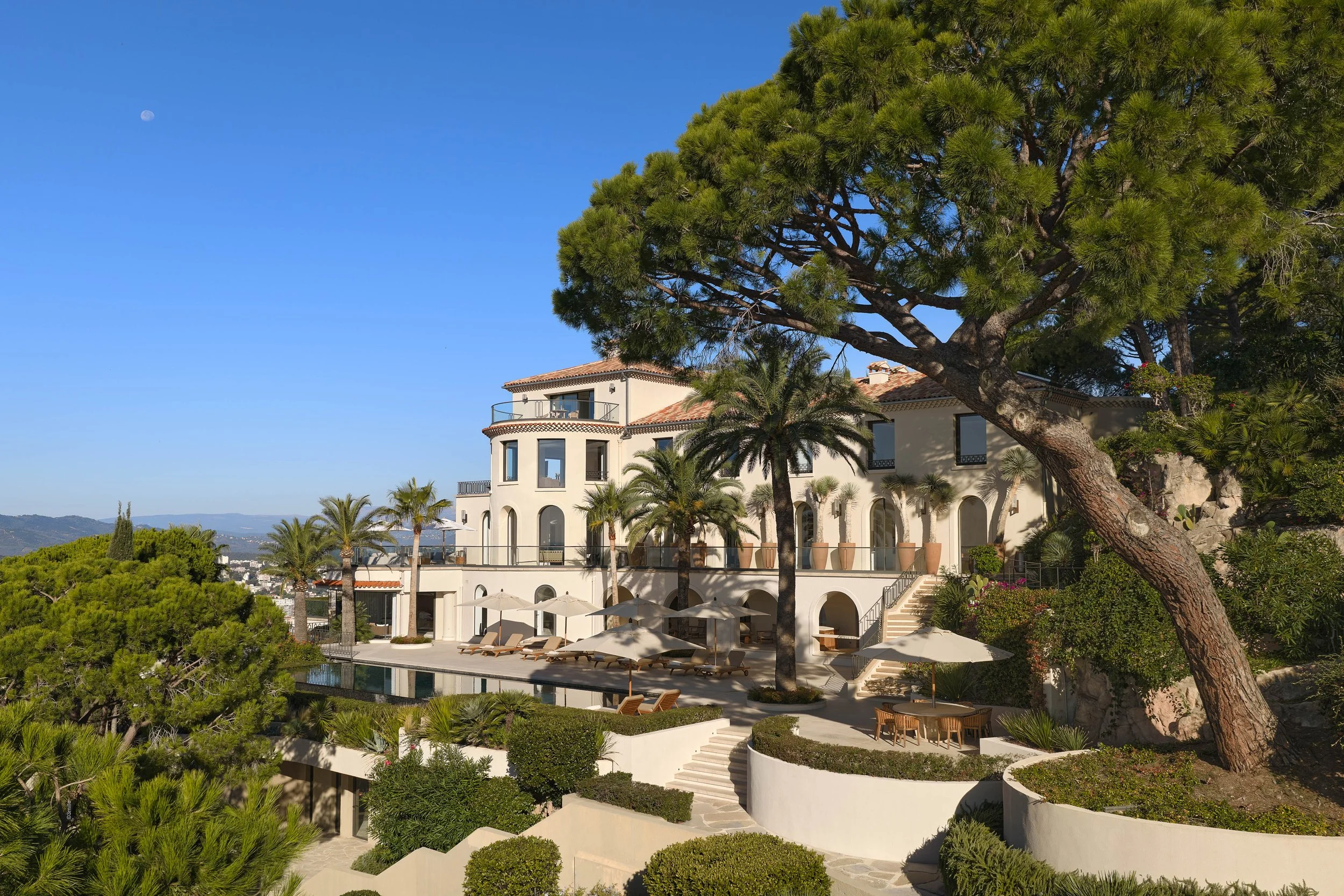 Villa de style méditerranéen avec plusieurs niveaux, entourée de jardins luxuriants, palmier et pins parasols.