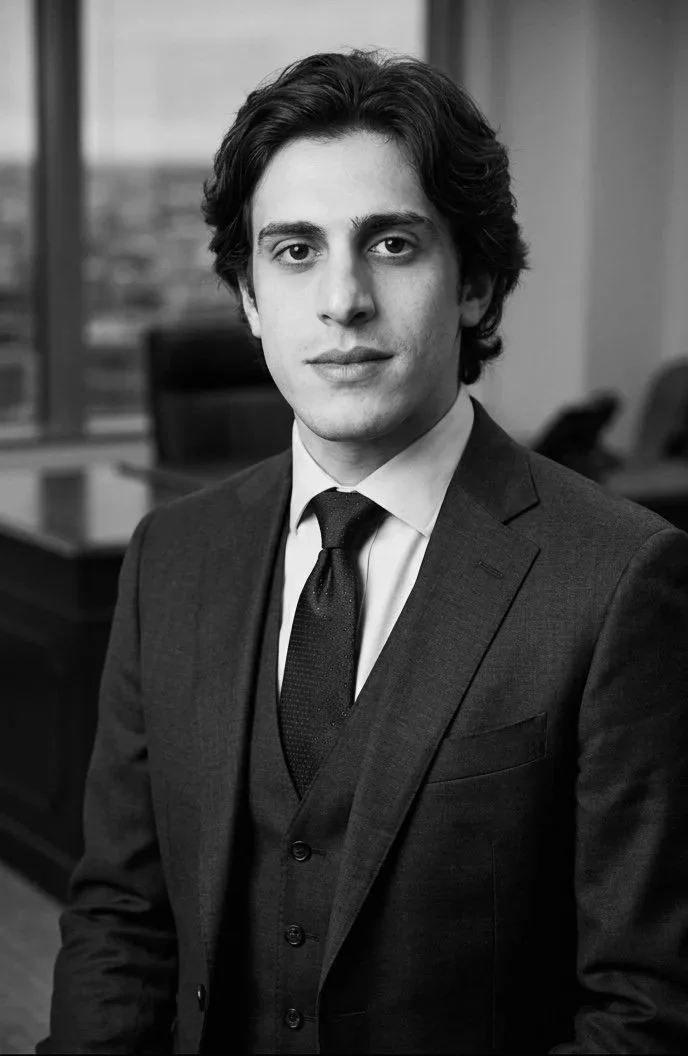 A young lawyer with medium-length dark hair, wearing a suit and tie, sitting in an office environment with large windows in the background.