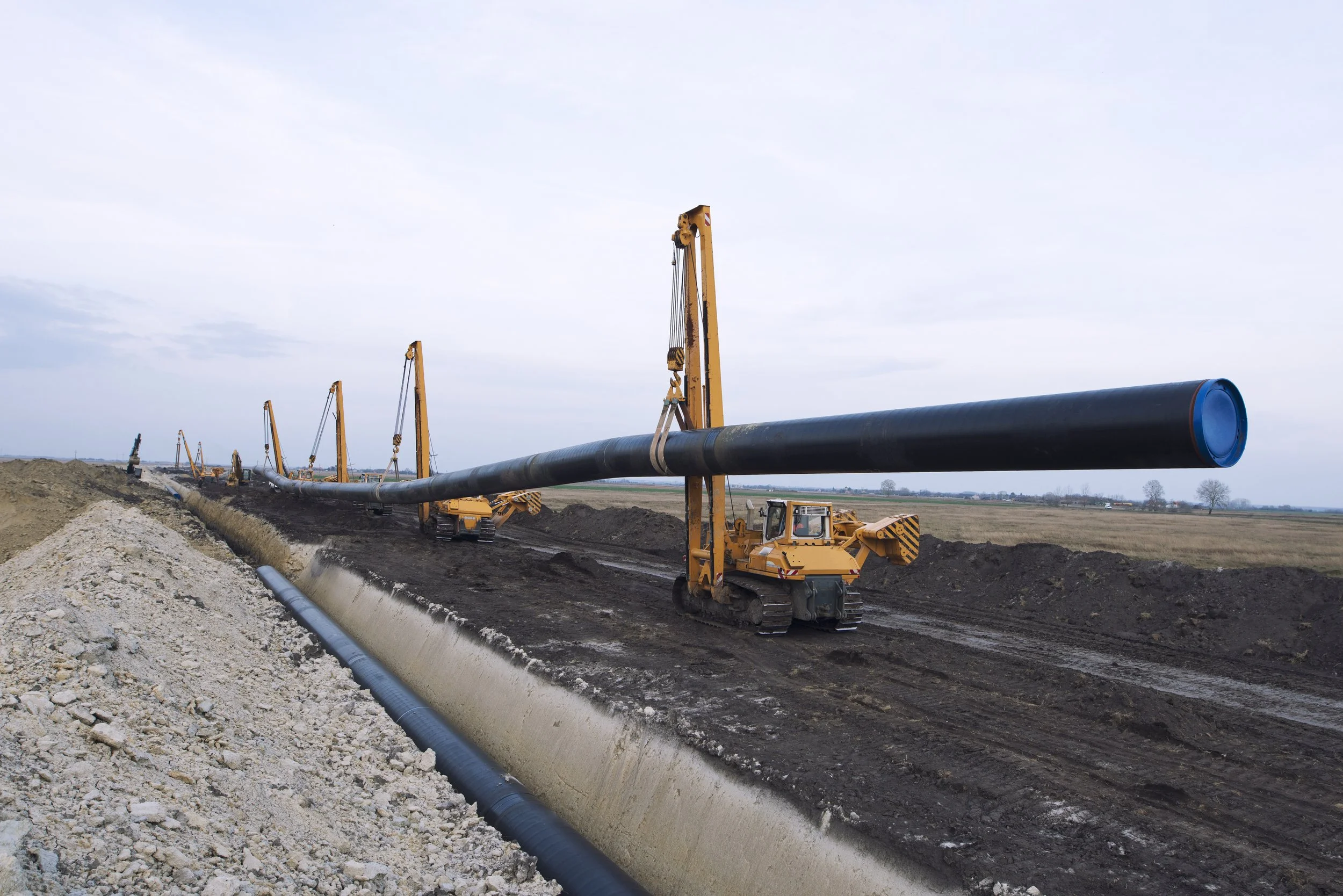 Construction site where large black pipes are being installed using yellow cranes on a dirt pathway, with a rural landscape in the background under an overcast sky.