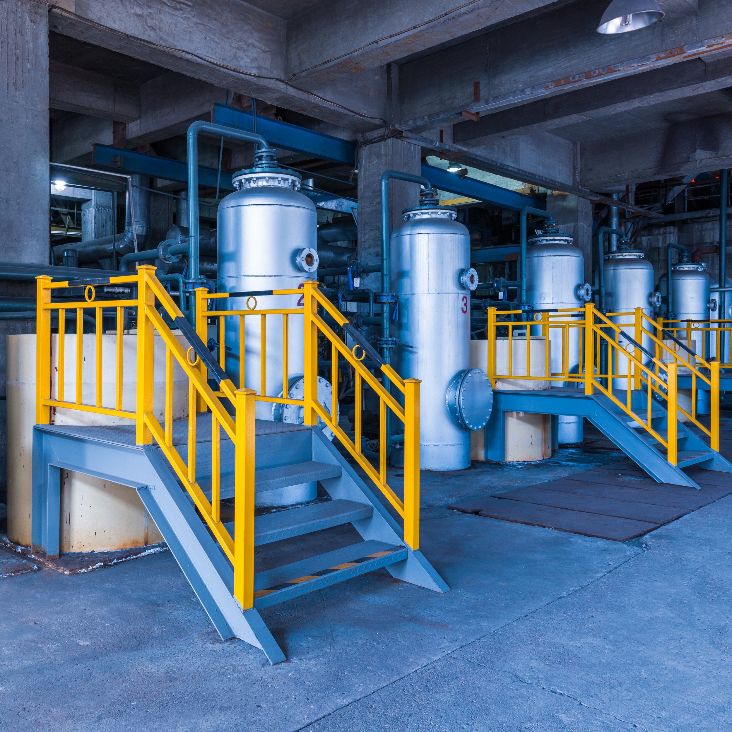 Industrial setting with four large silver tanks, yellow railings, and metal stairs, inside a concrete building.