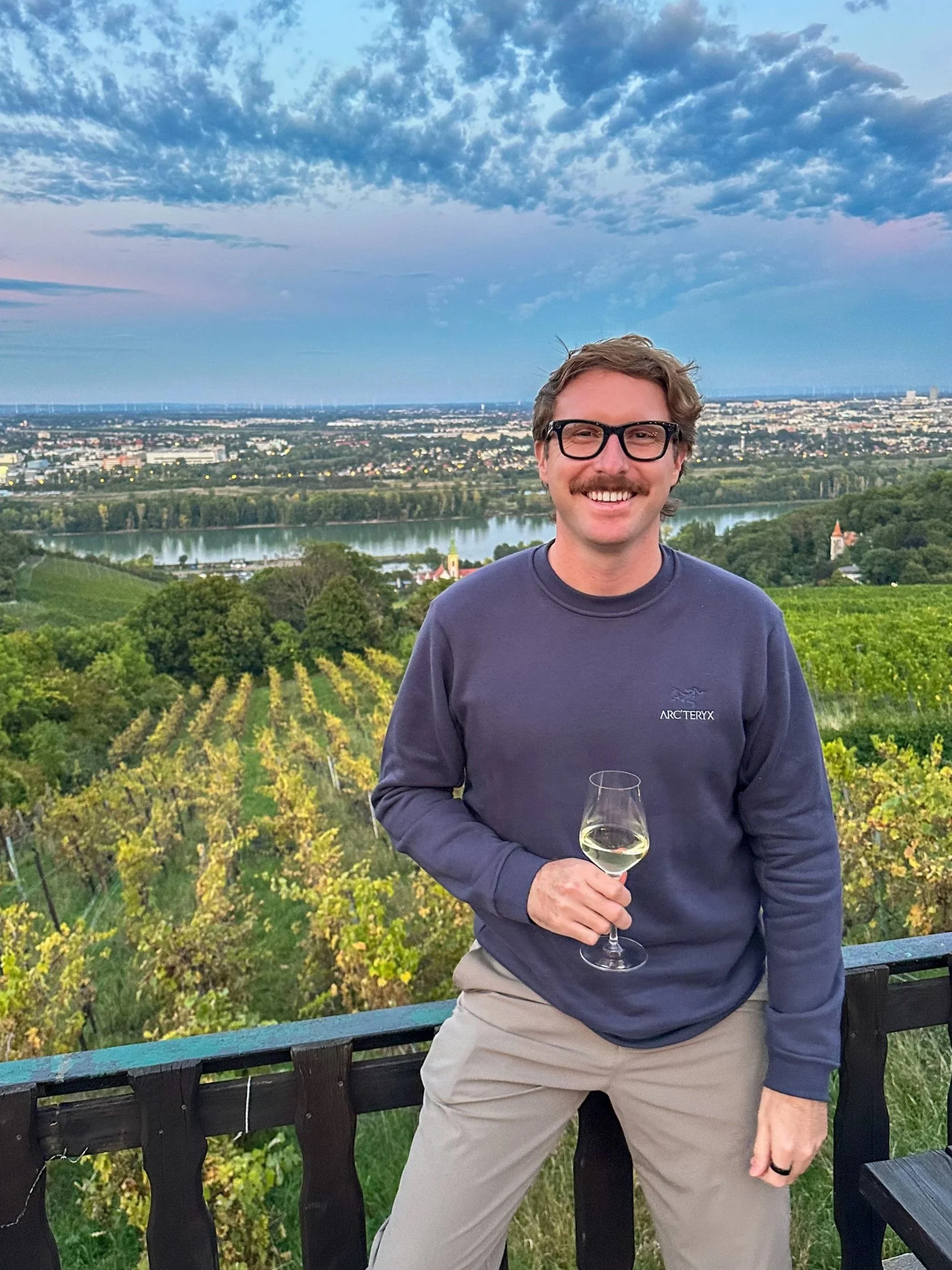 A man with glasses and a mustache wearing a navy sweatshirt and khaki pants, holding a glass of white wine, standing outdoors with a view of vineyards, a river, and a city skyline at dusk.