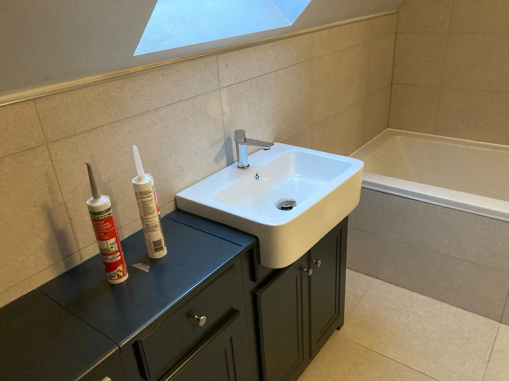 A bathroom with beige tiled walls and floor, featuring a small white sink with a silver faucet, black cabinet underneath, a black countertop with two tubes of adhesive or sealant, and a corner bathtub.