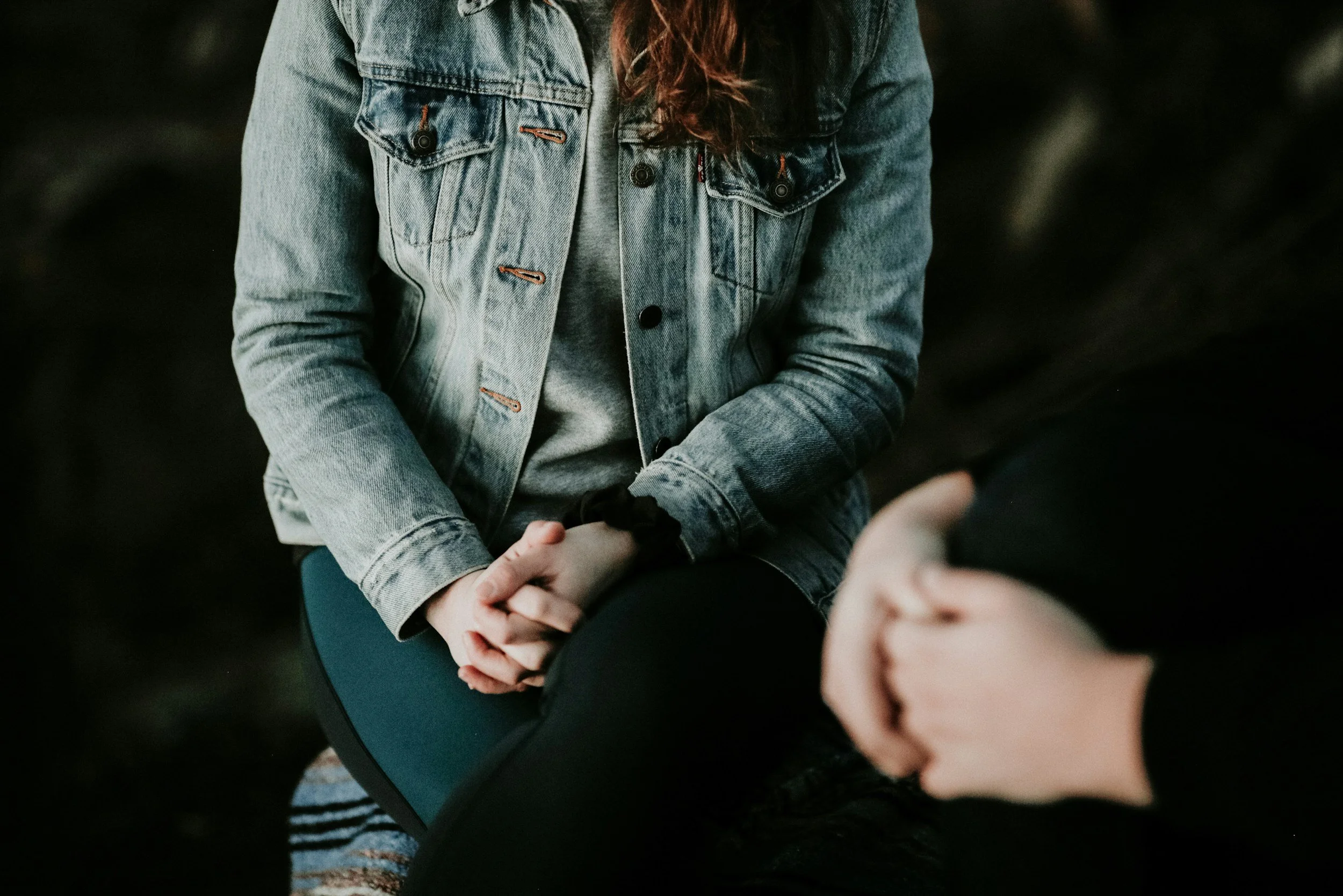 People sitting together, with one person wearing a denim jacket and the other person visible from the neck down, holding hands.
