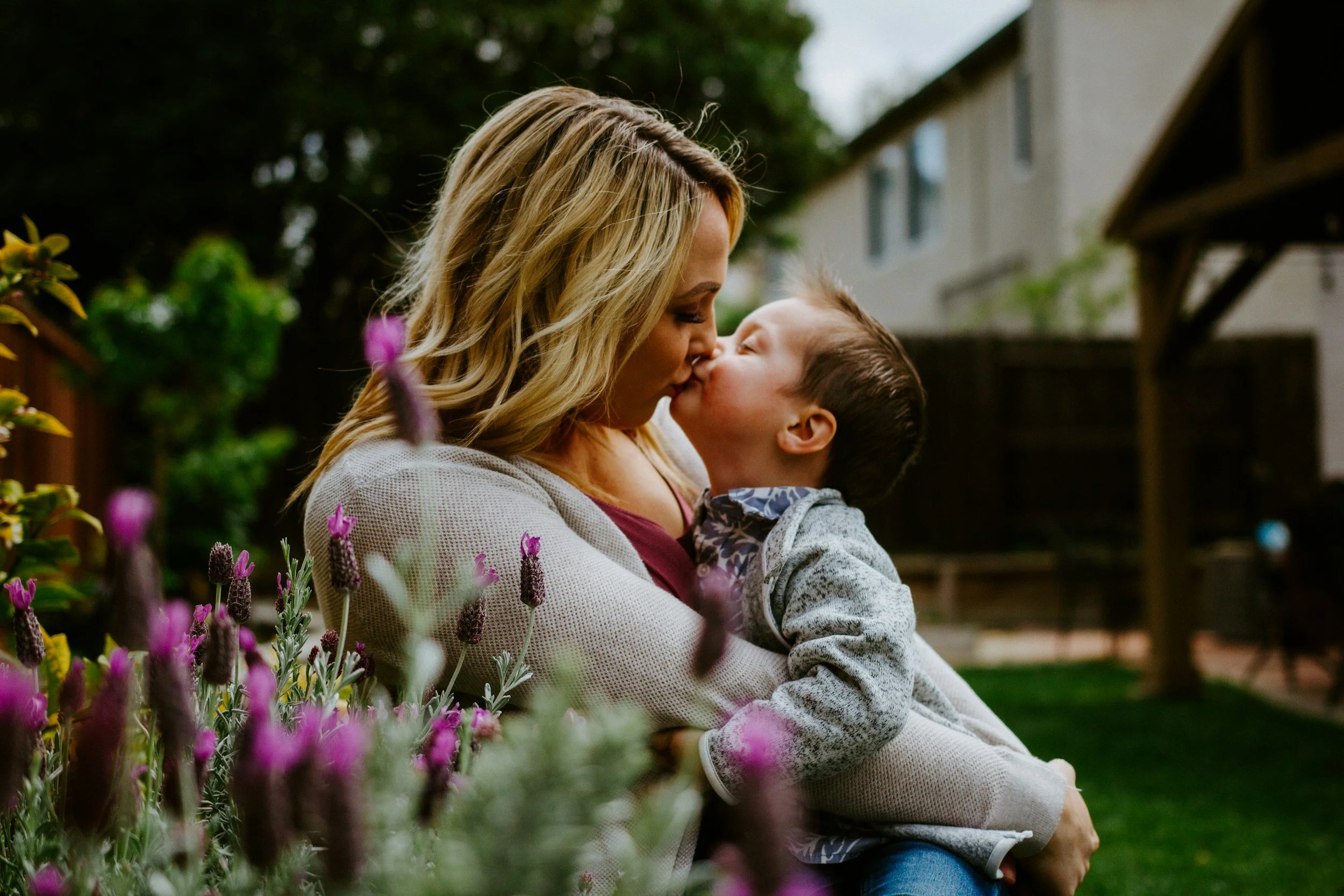 A woman with long blonde hair holding a young boy with dark hair and glasses, both sharing a kiss outdoors in a garden with purple flowers in the foreground.