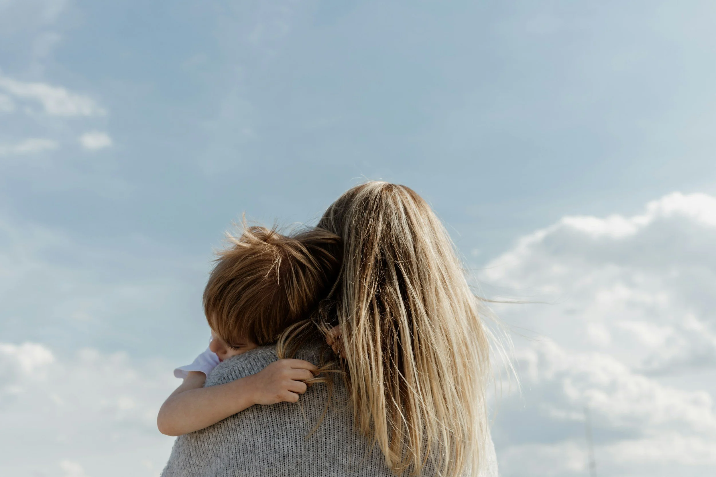A woman with long blonde hair hugging a child outdoors under a cloudy sky.