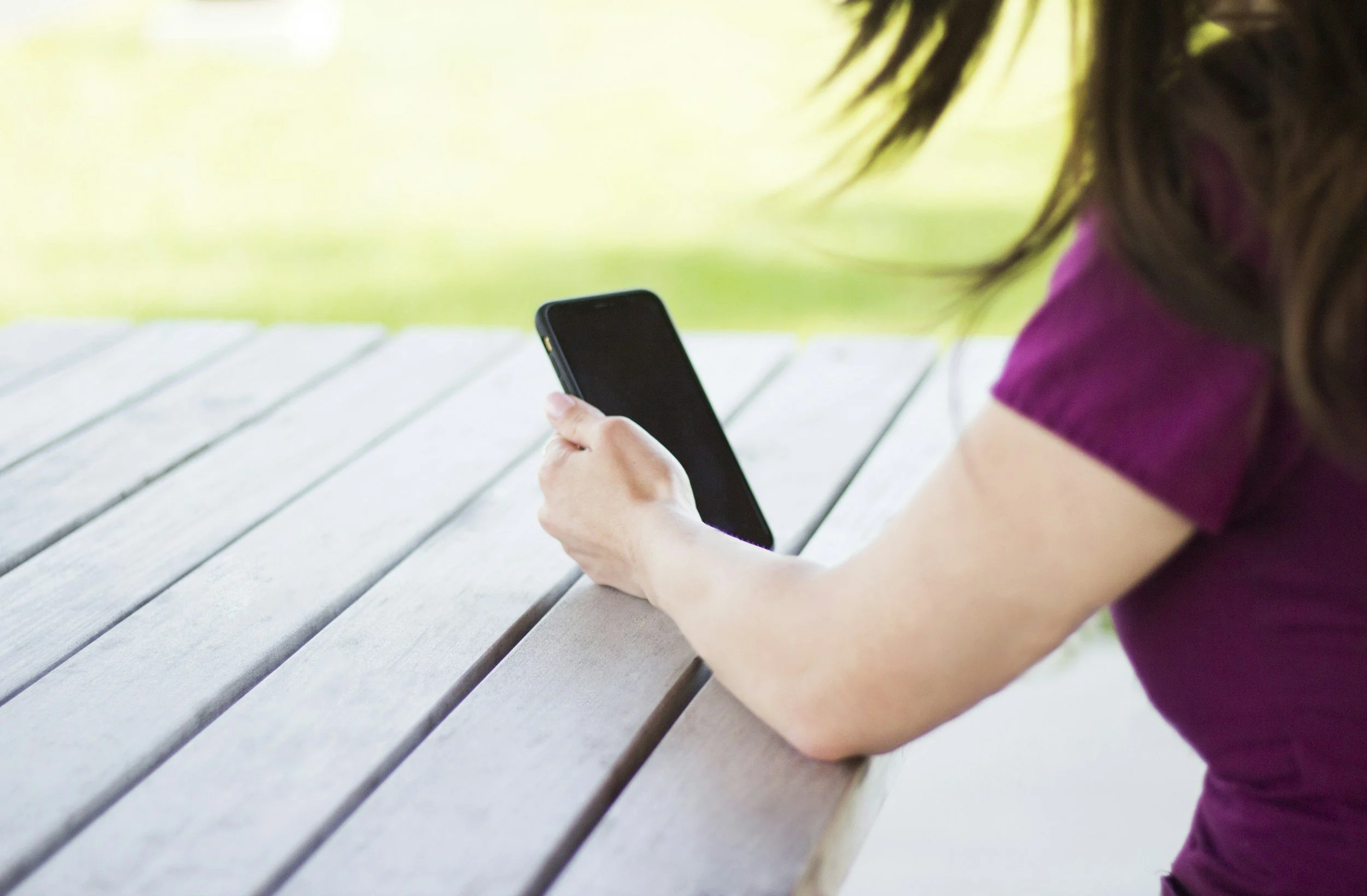 Person in a purple shirt sitting at an outdoor wooden table, holding a smartphone in their right hand, with grass and blurred greenery in the background.