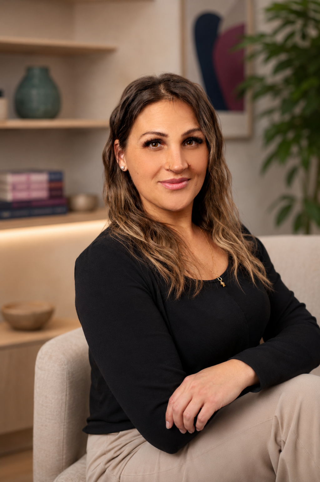 A woman with wavy brown hair, wearing a black long-sleeve top and beige pants, sitting on a beige chair in a living room with a smile, in front of a bookshelf with decorative items, a framed abstract painting, and a potted plant.